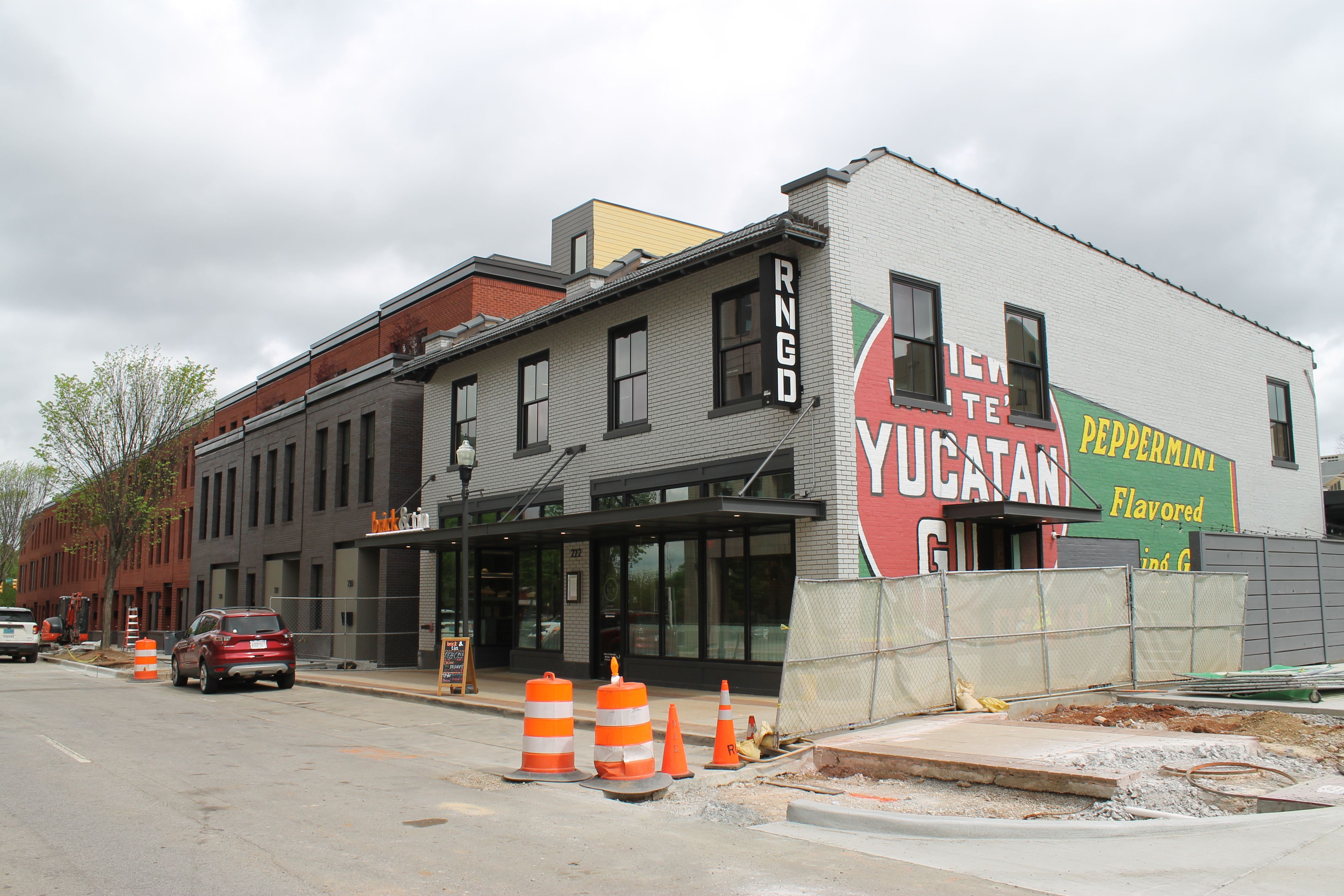 Street with a row of brick storefronts under construction, orange cones and fencing along the sidewalk. A large mural on the side reads "YUCATAN" and "PEPPERMINT" against an overcast sky.