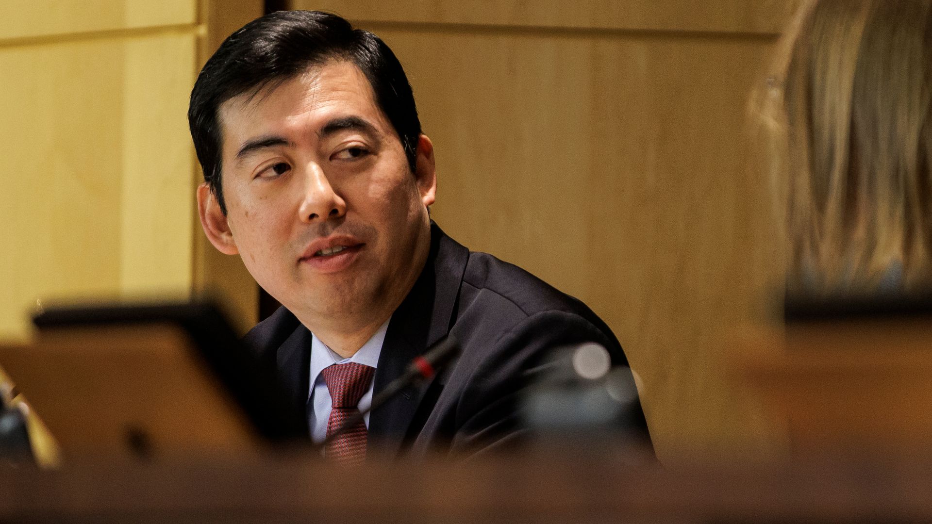 A man with dark hair and a dark grey suit in an official looking meeting, with lots of hardwood around.