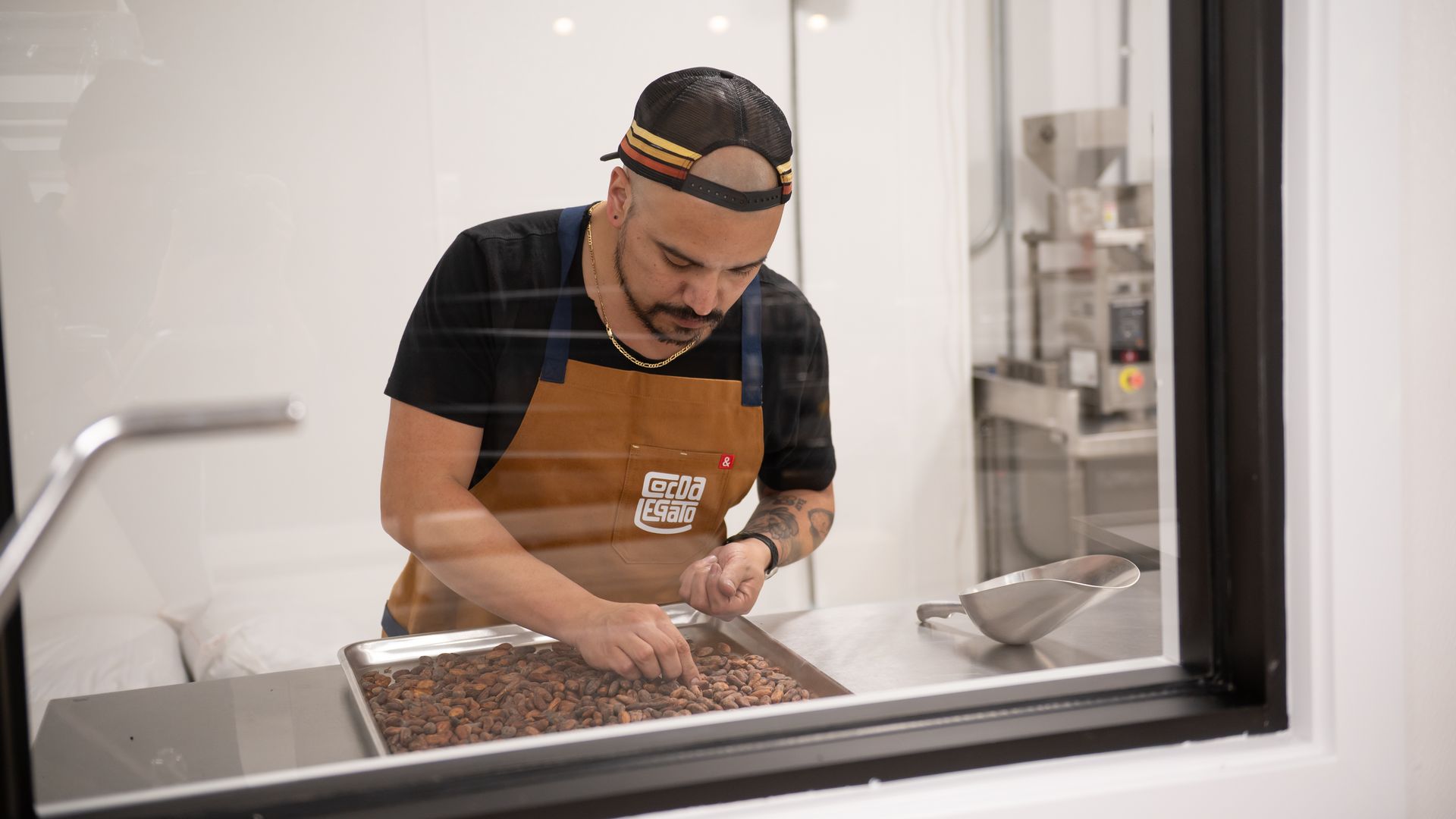 Aaron Lindstrom wears a brown apron and sorts cacao beans on a tray behind a window.