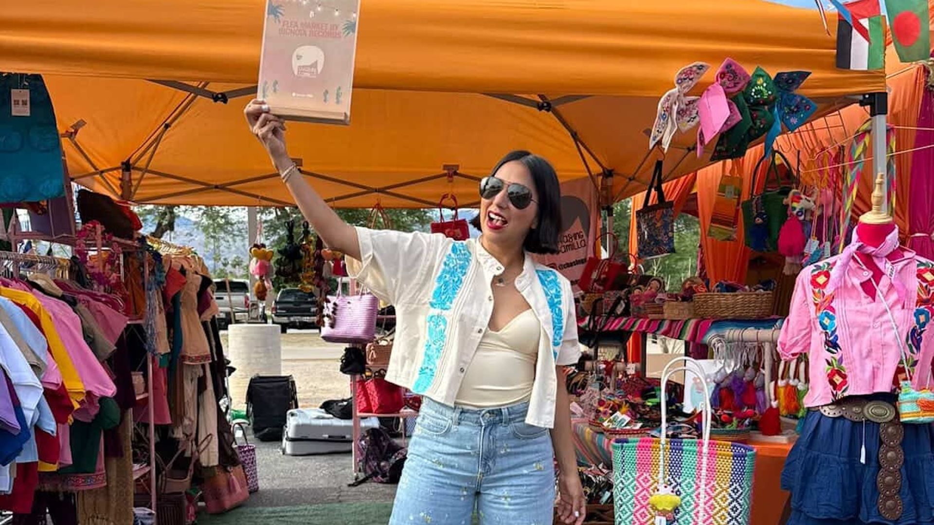 Woman in sunglasses, white top and denim shorts poses under an orange market canopy, holding a certificate aloft. Colorful clothes, bags, and flags surround her; an orange tote reads "nadie me cae bien."