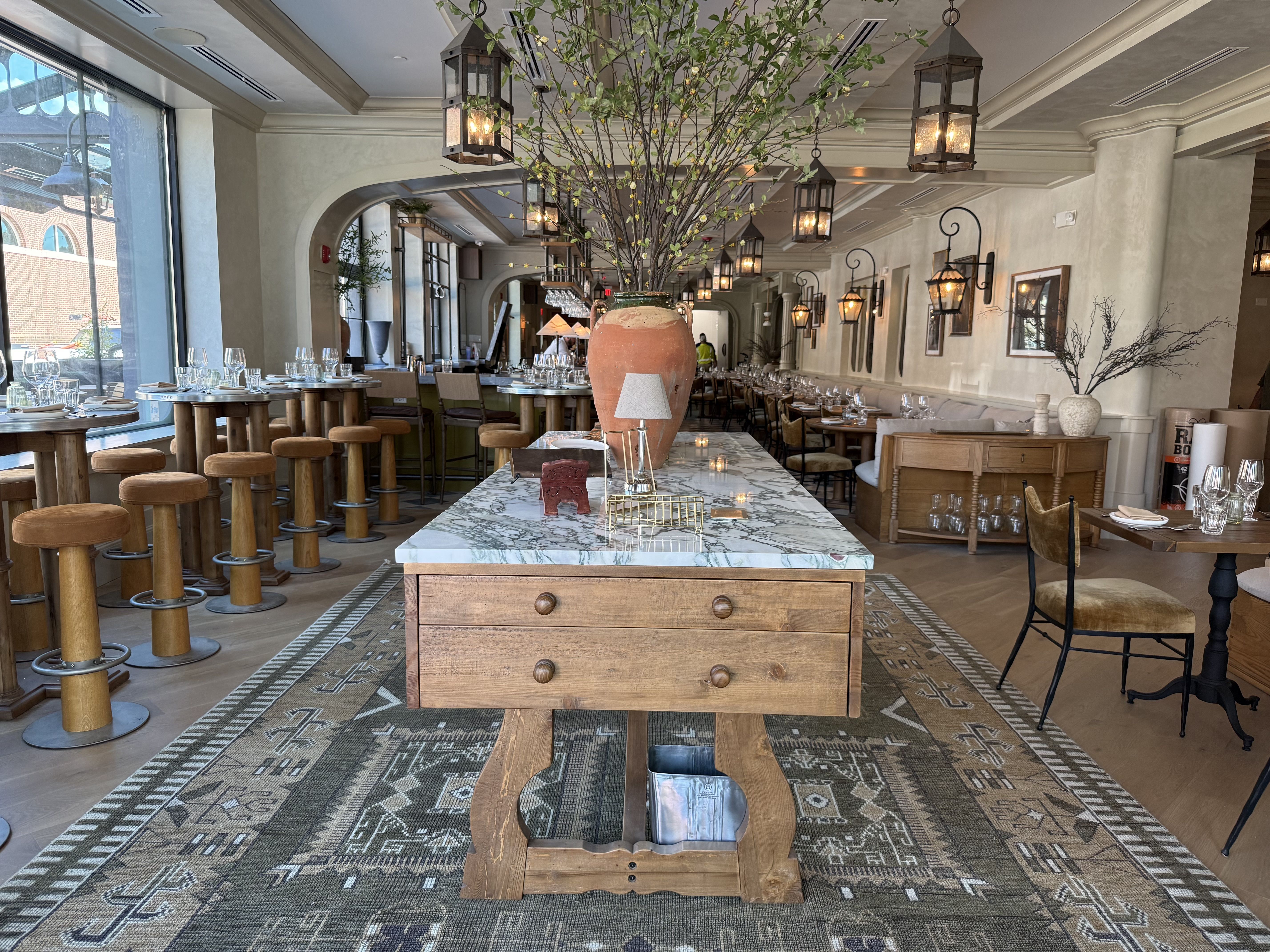 Bright restaurant interior with a marble central island, a large terracotta vase of branches, suspended lanterns, wooden stools at high tables, arched doorways, and large windows.