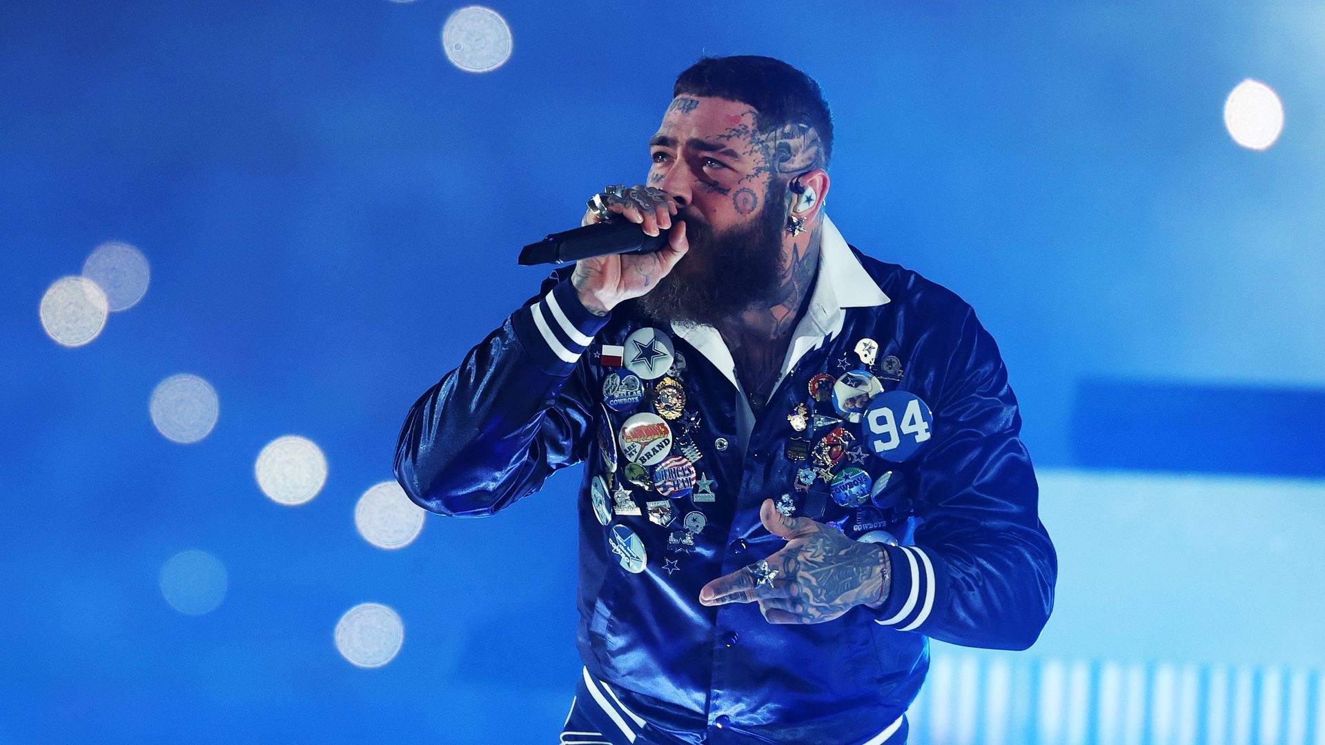Bearded tattooed man wearing a blue satin jacket with many pins and patches, holding a microphone while performing against a blue background with light orbs.