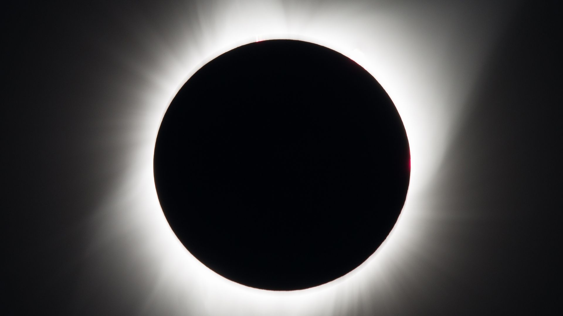 Close-up of a solar eclipse: a dark Moon covers the Sun, with a bright white corona radiating outward and a faint red rim on the right edge against a dark sky.
