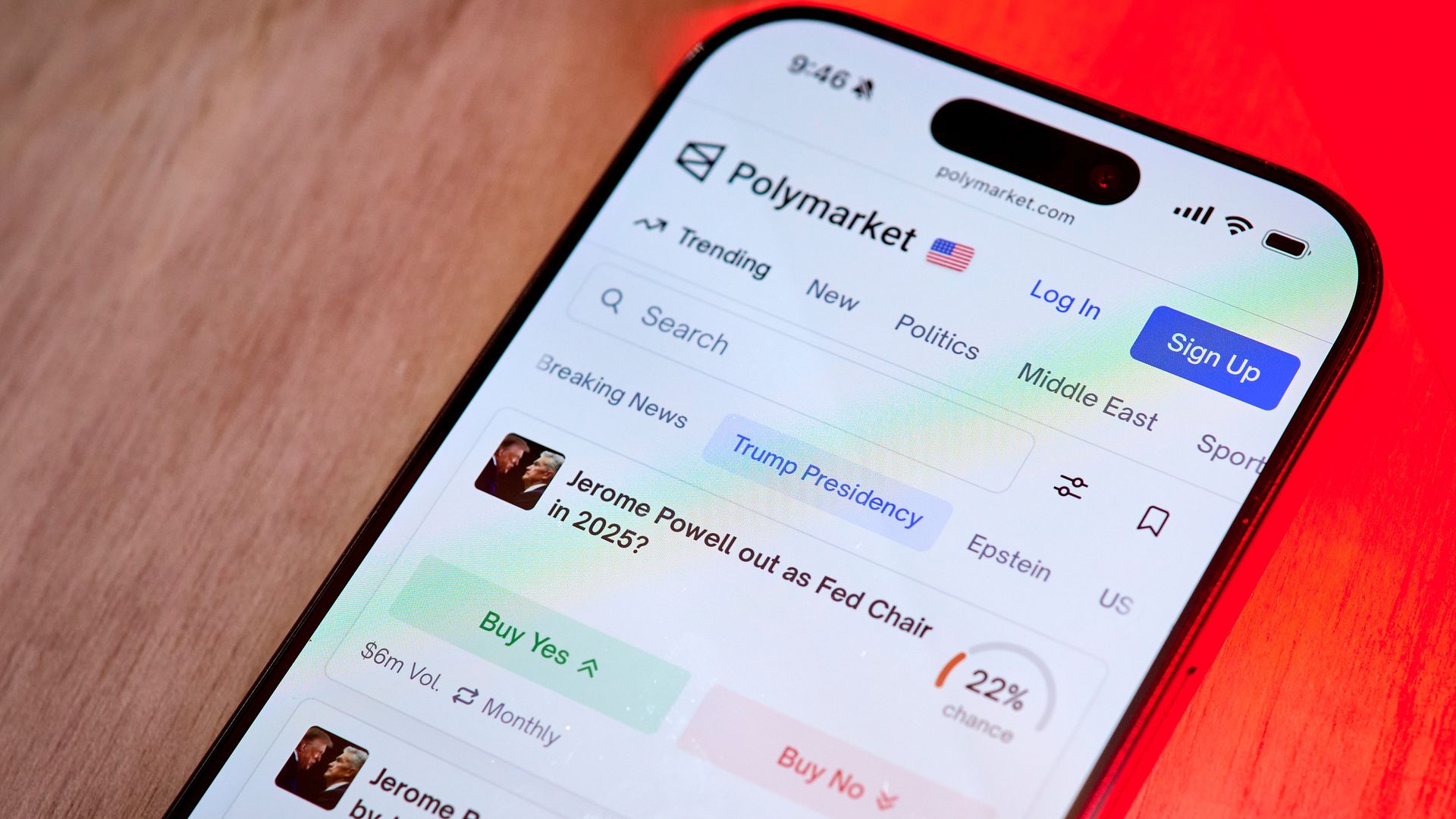 Smartphone screen displaying Polymarket website with predictions about Jerome Powell's Fed Chair status in 2025 and by July 31, featuring green and red buy buttons on a wood and red surface.