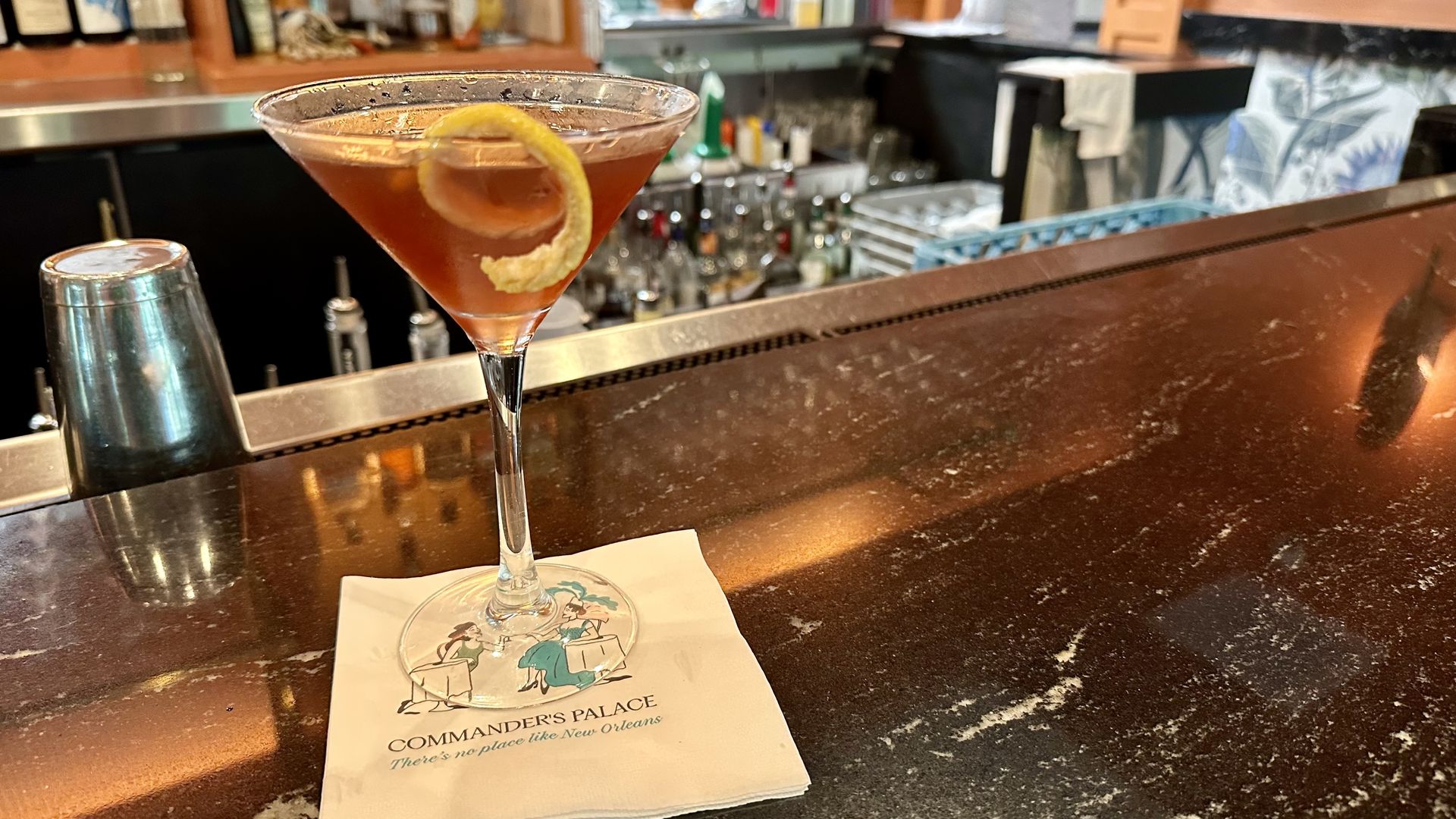 A purple cocktail in a martini glass garnished with a lemon twist sits on a napkin on a copper bar, with a busy back bar of bottles and equipment in the blurred background.
