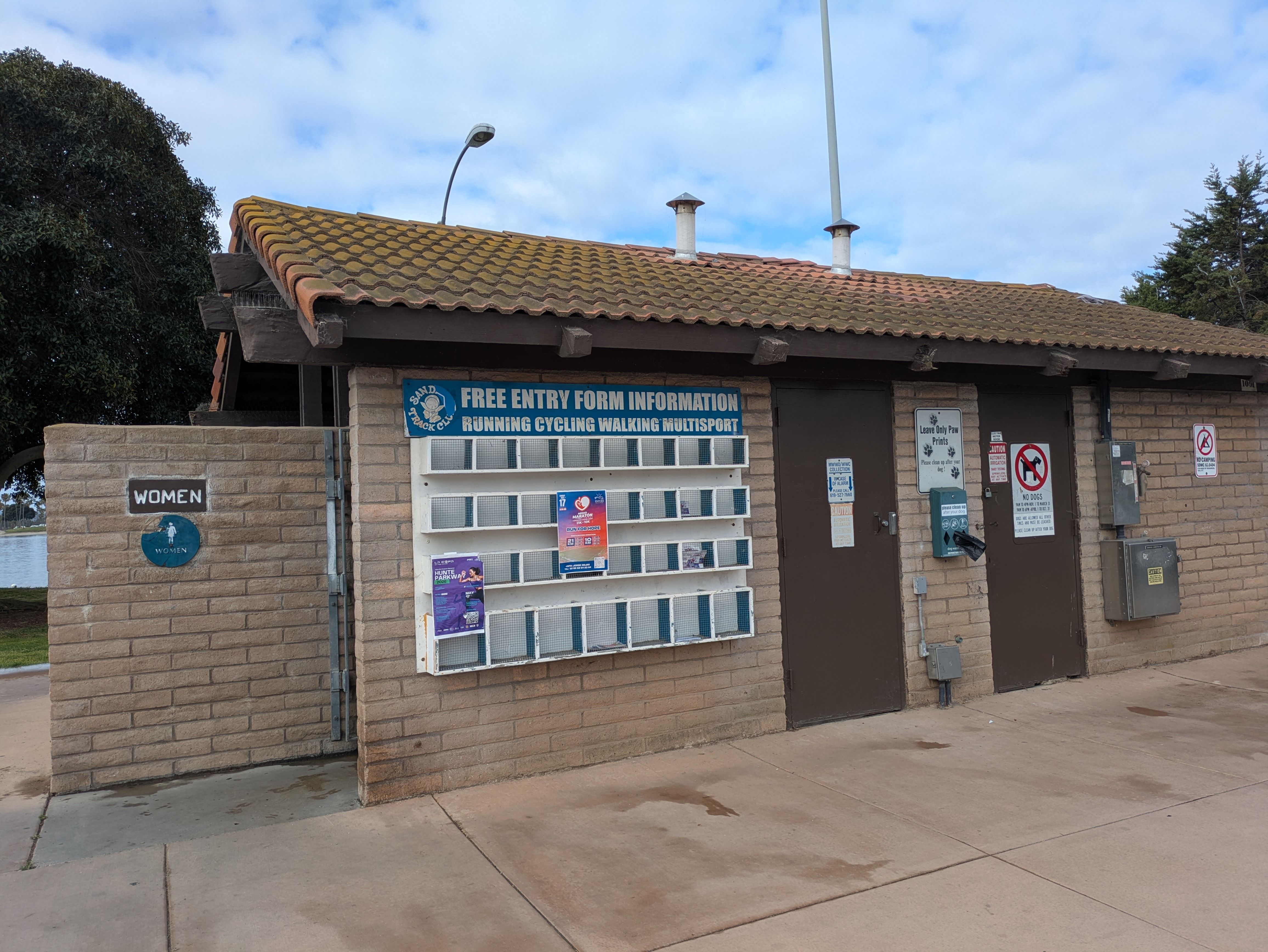Small brick information booth with a brown tiled roof, two chimneys, and a curved street lamp. A blue poster reads FREE ENTRY above the display; left wall shows a WOMEN sign. Clear sky.