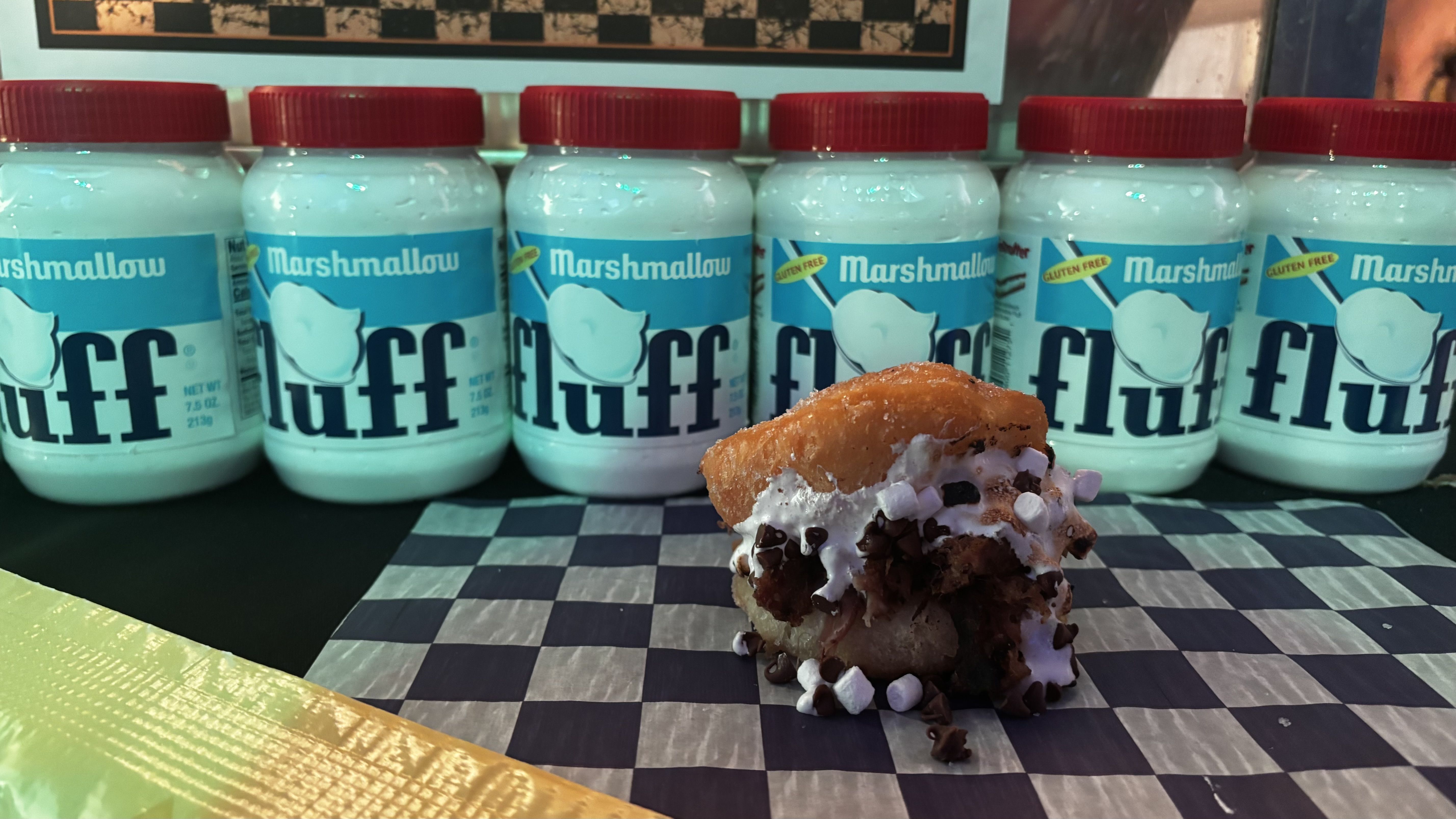 Teal Marshmallow Fluff jars with red lids lined up behind a stack of ice cream sandwiches topped with whipped cream and chocolate chips on a black-and-white checkered table.