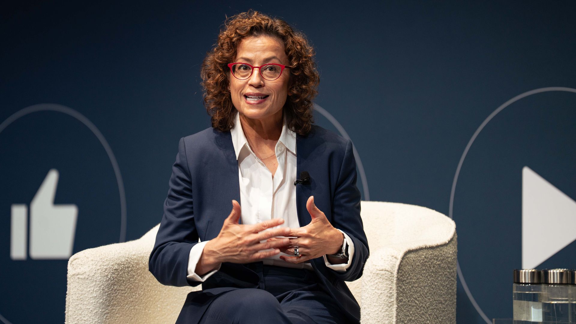 FCC commissioner Anna Gomez speaks from a white armchair, wearing a navy blazer and white blouse, with social media like and play icons in the background.