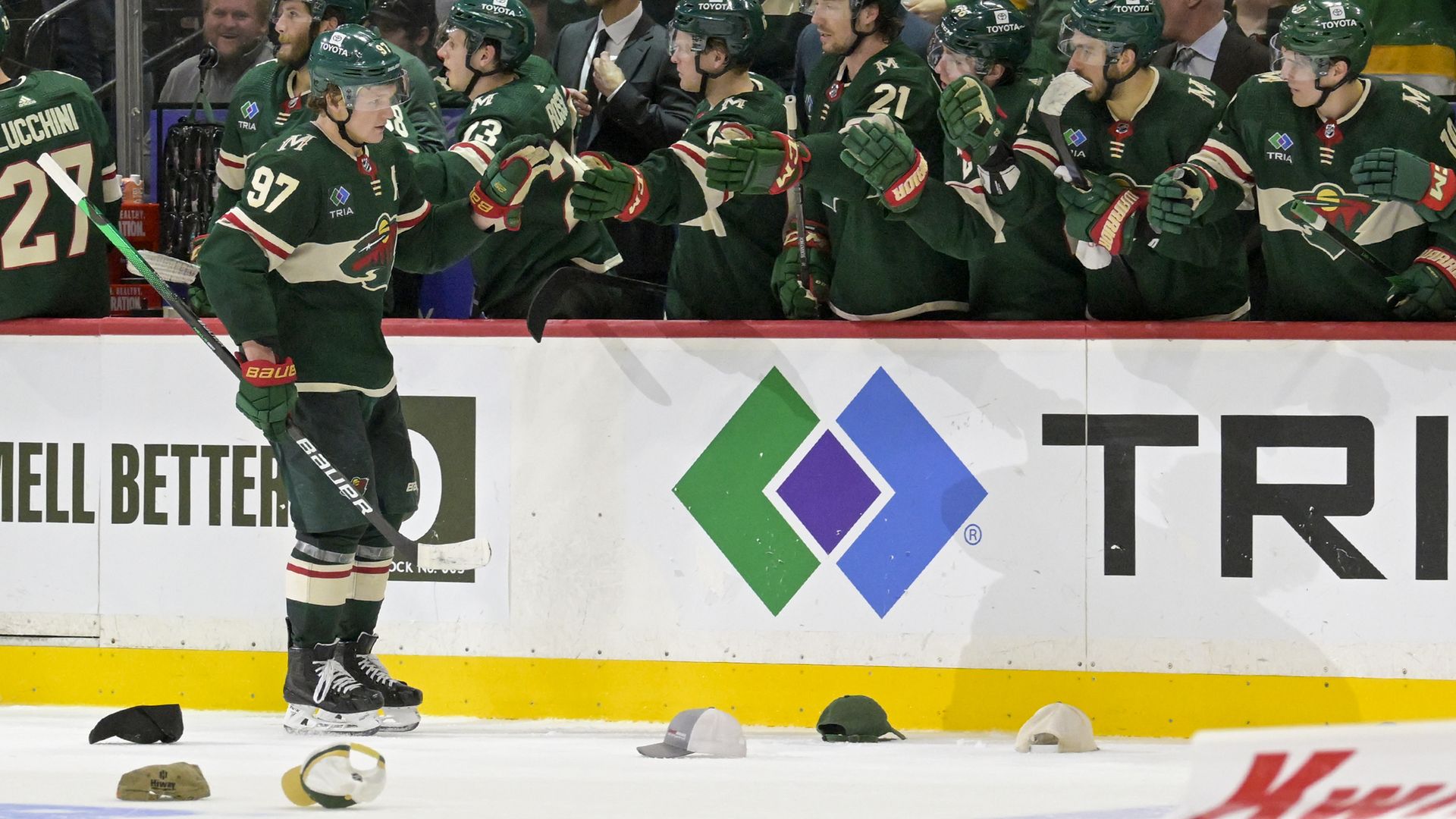 A hockey player in a dark green jersey with cream-colored trim skates along his team's bench, fist-bumping his teammates over the boards. Hats from the player's "hat trick" third goal of the game litter the ice.