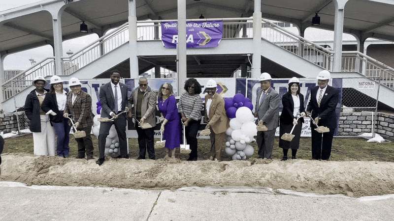 Group of people, some in hard hats, at a groundbreaking ceremony holding shovels with dirt. Purple and white balloons and a banner reading "Rolling into the Future" behind them.