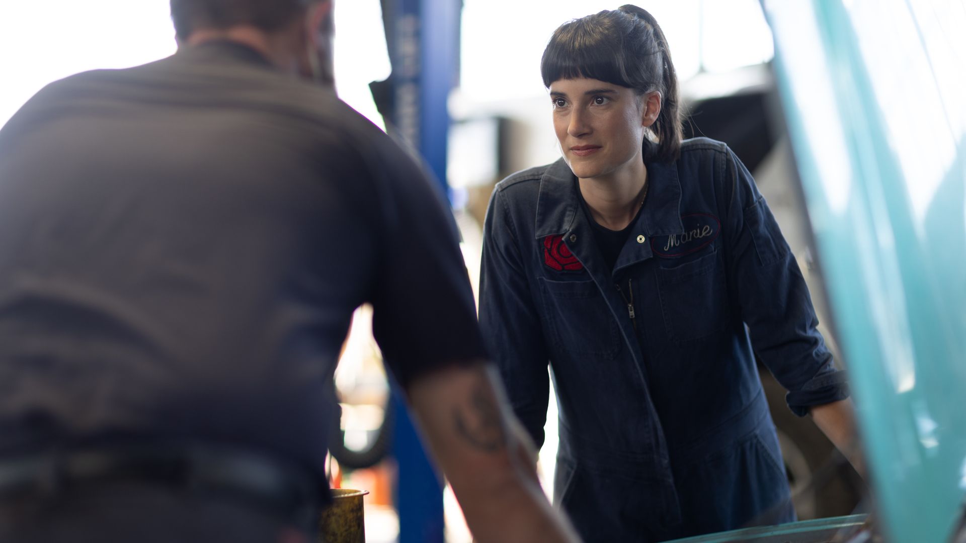 A woman in coveralls rests her arms on the open hood of a car, while speaking to someone whose back is to the camera. 