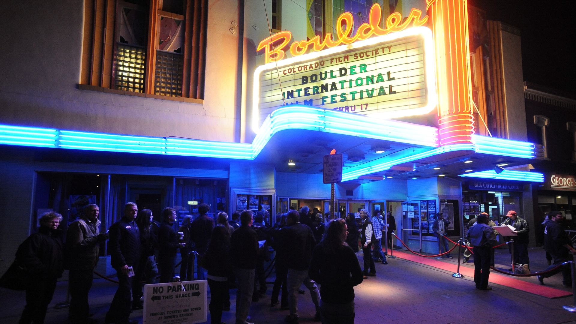 Night scene outside the Boulder Theater with neon signage reading "Boulder International Film Festival" and bright blue marquee lights. A queue of people waits at the entrance beneath a red rope barrier.