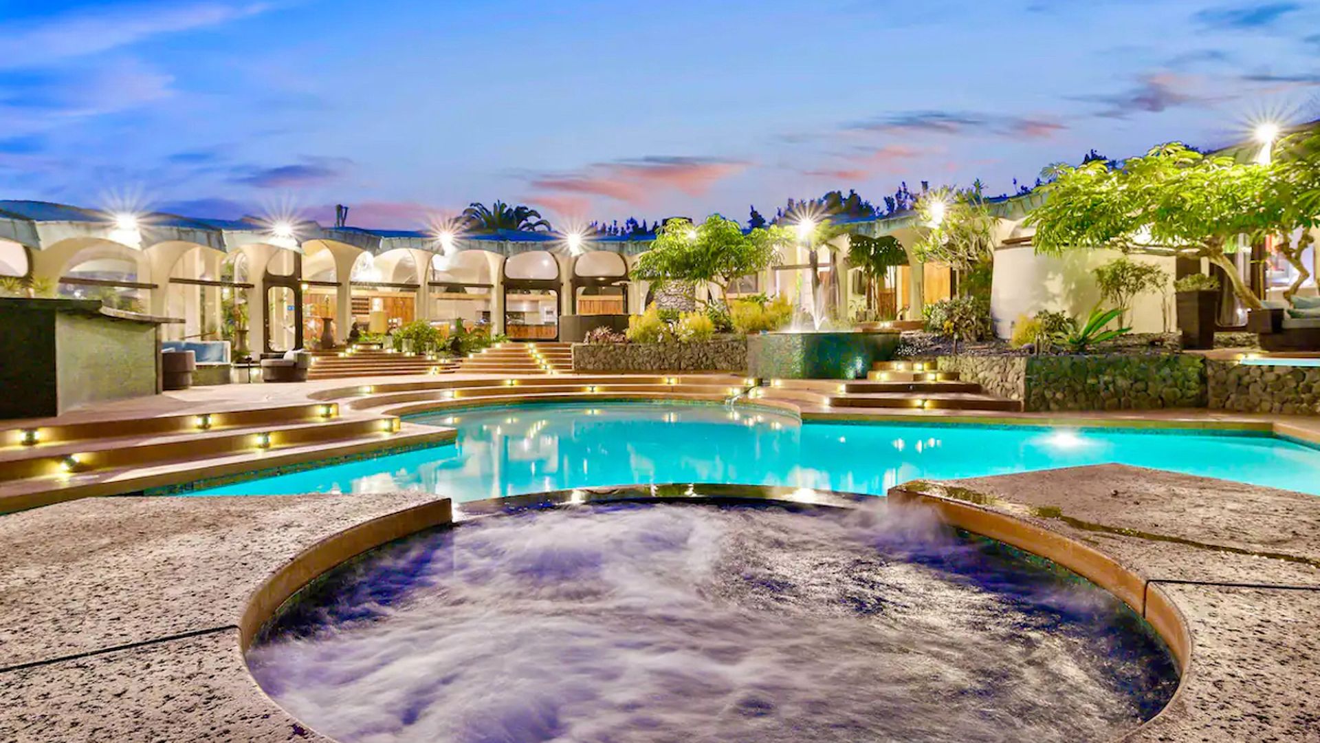 A hot tub and resort-style pool are featured in front of an expansive airbnb home at sunset. 