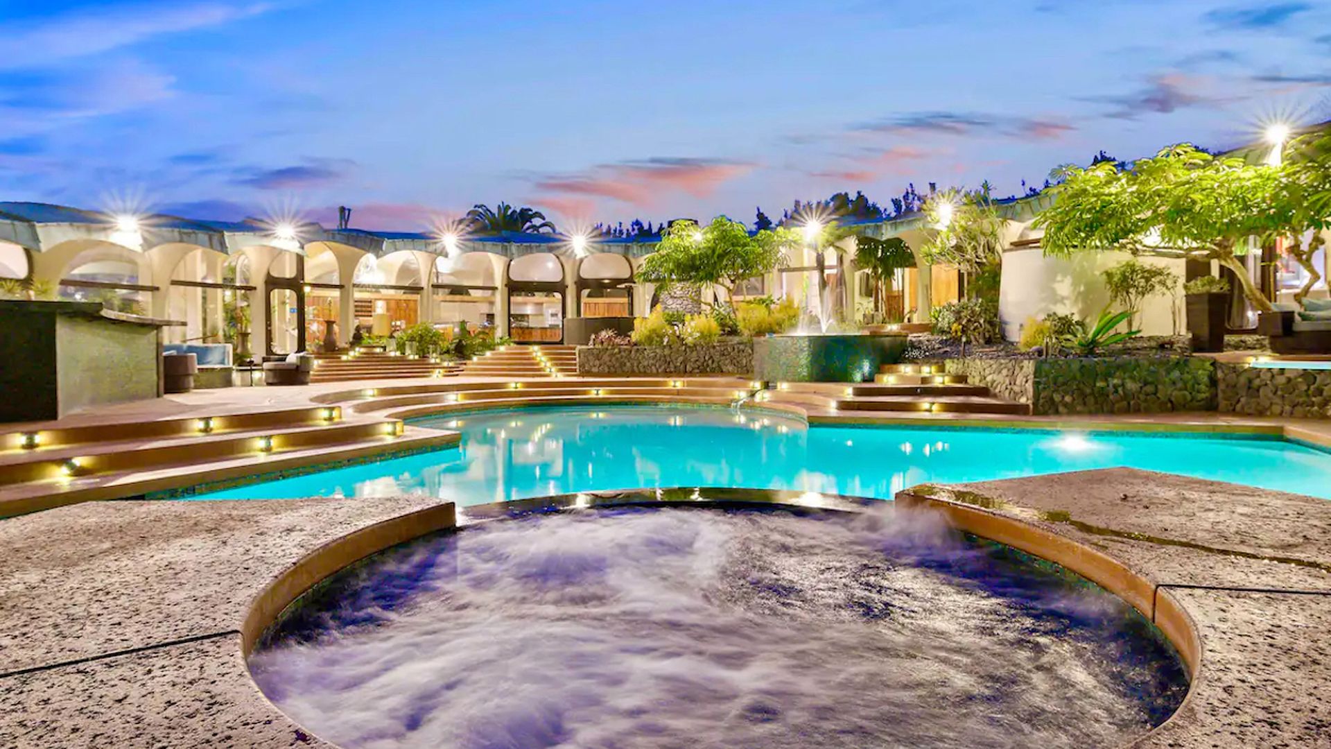 A hot tub and resort-style pool are featured in front of an expansive airbnb home at sunset.