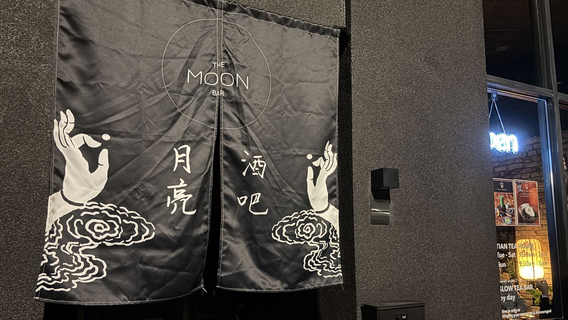 Dark textured wall with two black banners showing a circle logo reading "THE MOON BAR" and white wave/cloud art at the bottom; a window with blue neon and posters sits to the right.