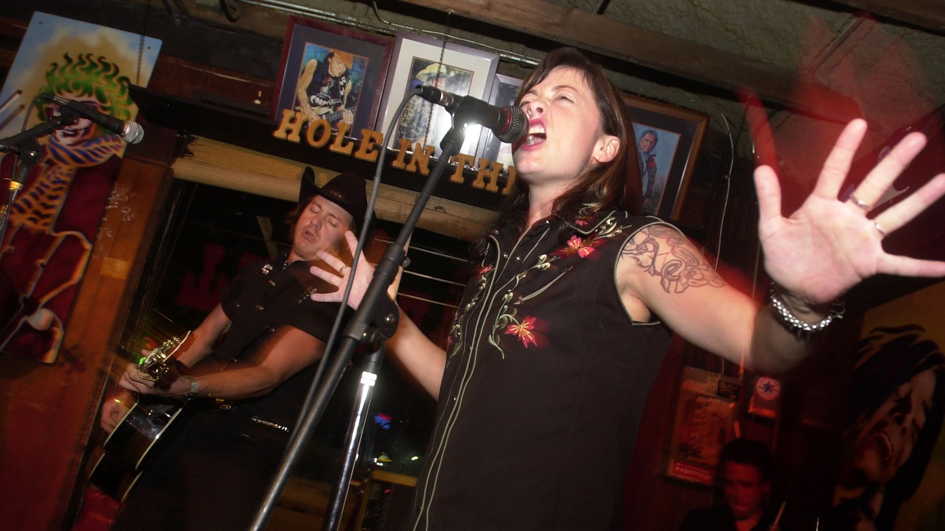 Female singer with a tattooed arm, sleeveless black shirt, belts into a microphone with mouth open; her hand is raised toward the camera; a guitarist in black plays in a cozy bar behind her.