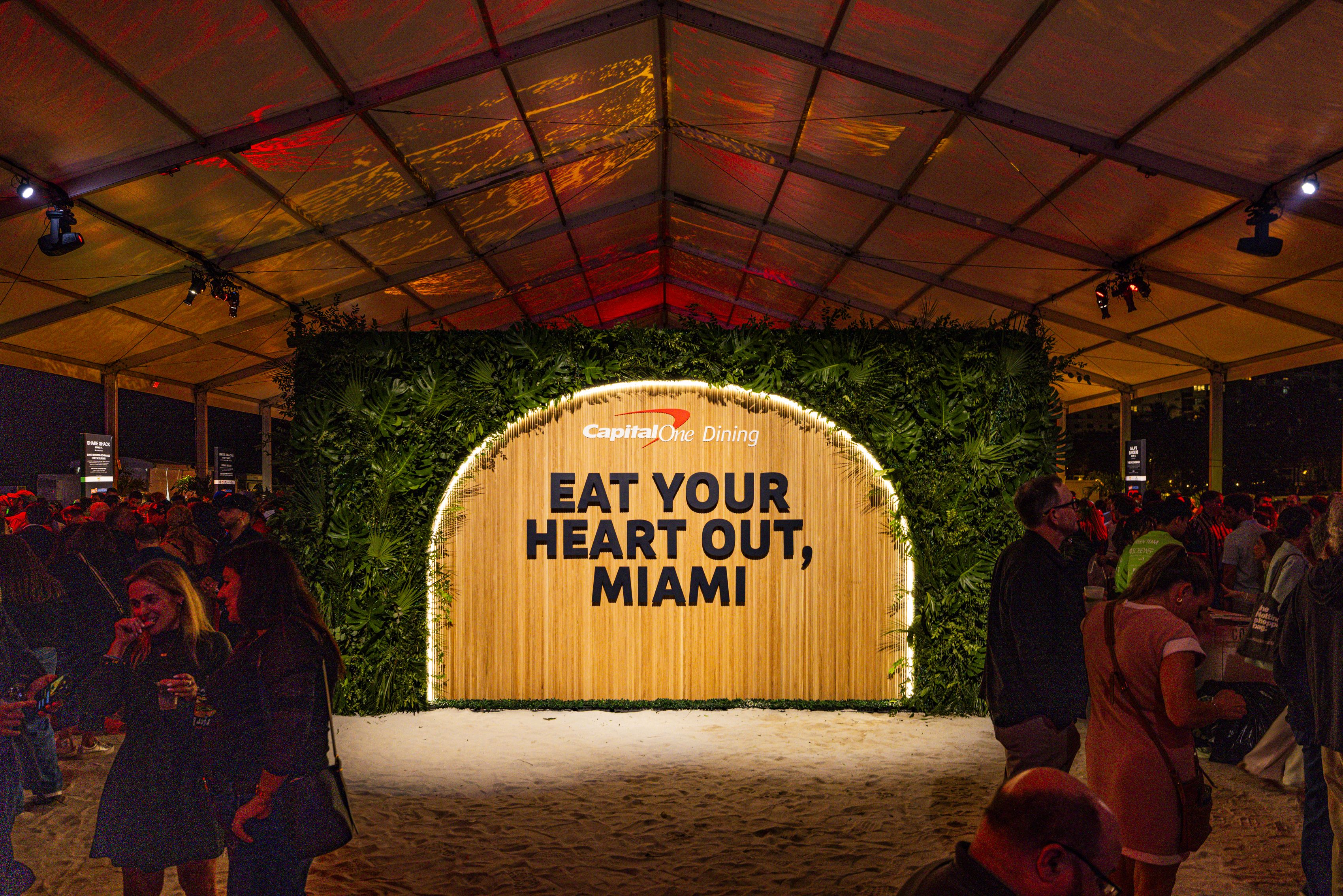 Indoor event tent with crowd, illuminated wooden sign framed by greenery reading Capital One Dining 'EAT YOUR HEART OUT, MIAMI' against sand floor and red lighting overhead.