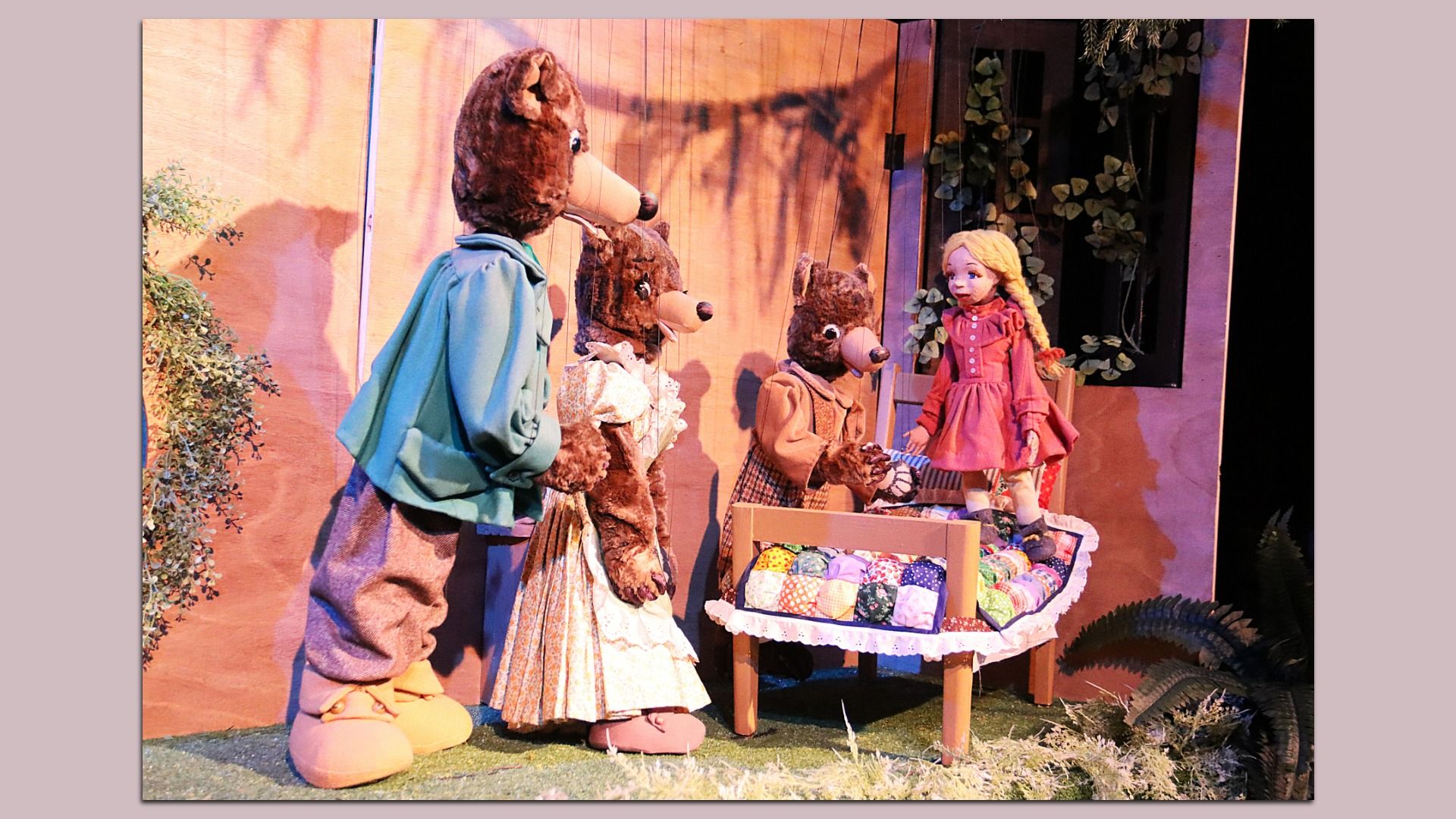 Stage puppet scene of Goldilocks and the Bears: a tall brown bear in a teal shirt speaks to a blonde girl in a pink dress with braids; two smaller bears stand nearby by a patchwork quilted bed under a wooden backdrop with vines.
