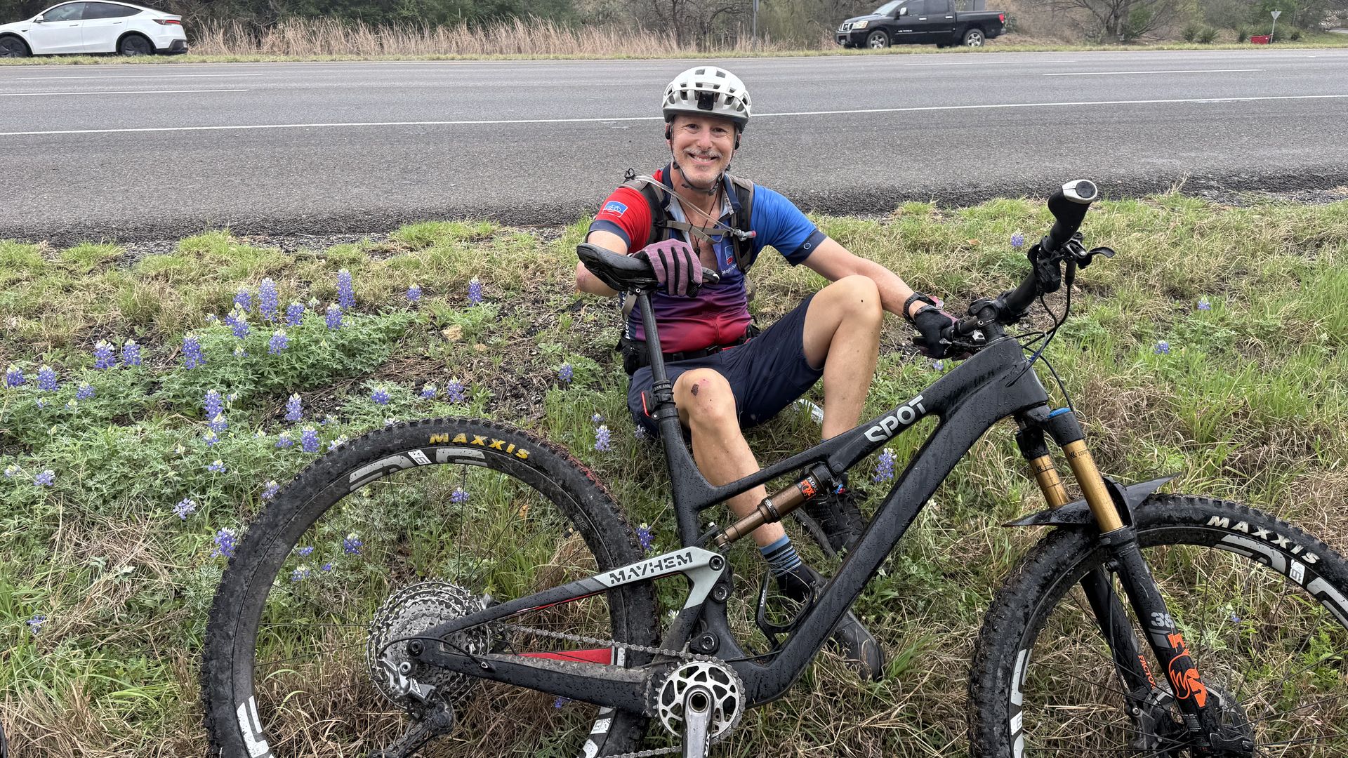 Smiling cyclist in a white helmet and red/blue jersey sits on a muddy black SPOT mountain bike by a grassy roadside, purple wildflowers in the foreground, and a road with cars in the background.