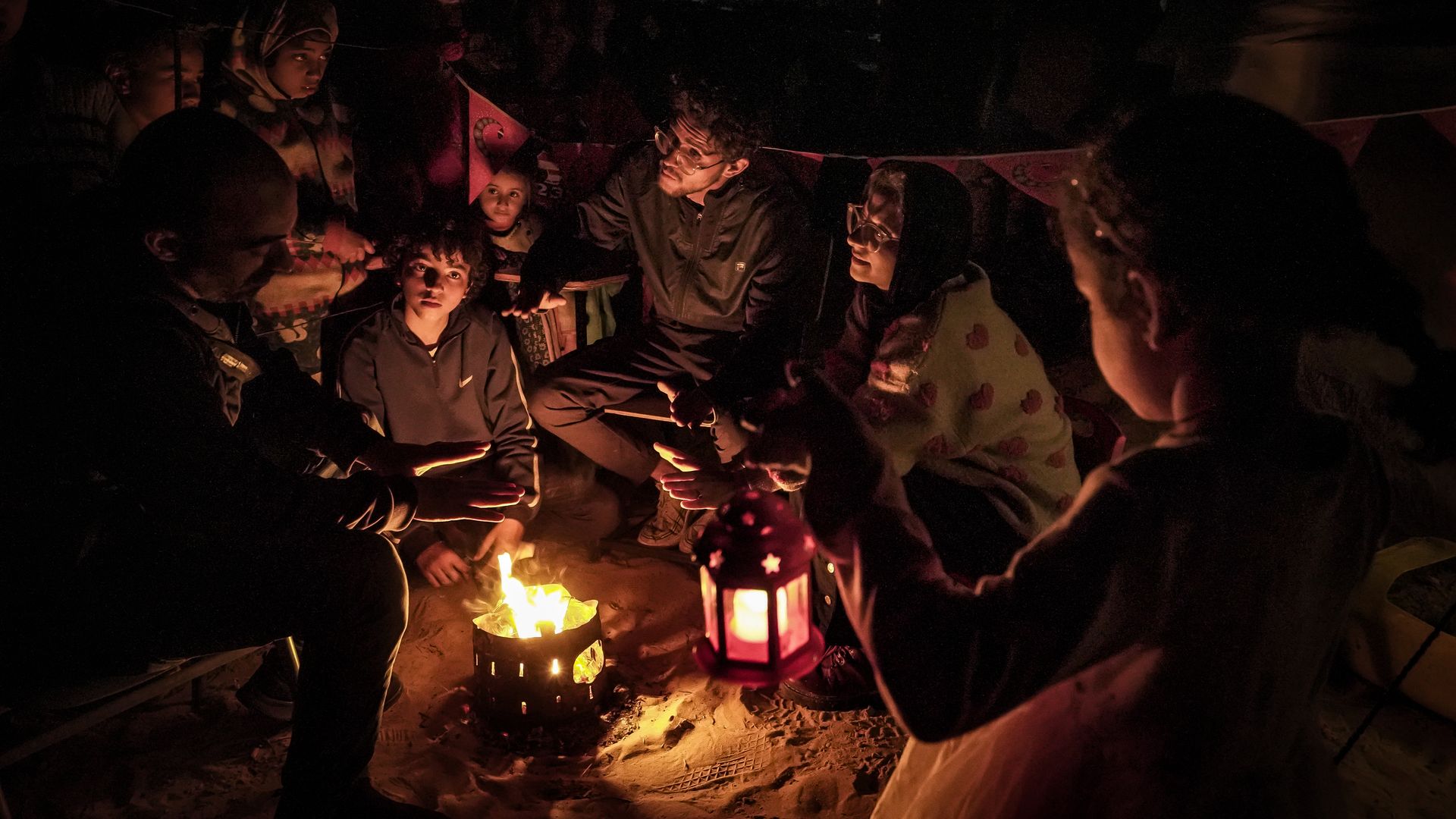 A young child is holding a lantern near a group of Palestinians people huddled around a fire. 