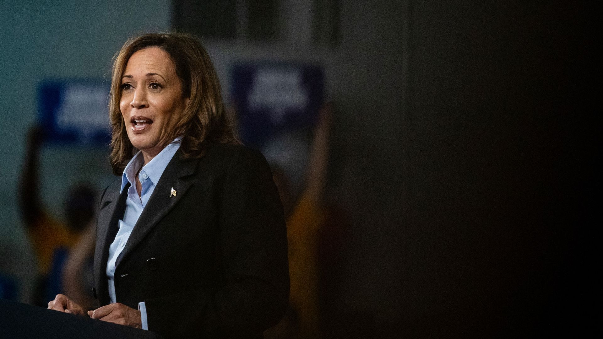 Vice President Kamala Harris stands at a lectern wearing a dark jacket and blue shirt 
