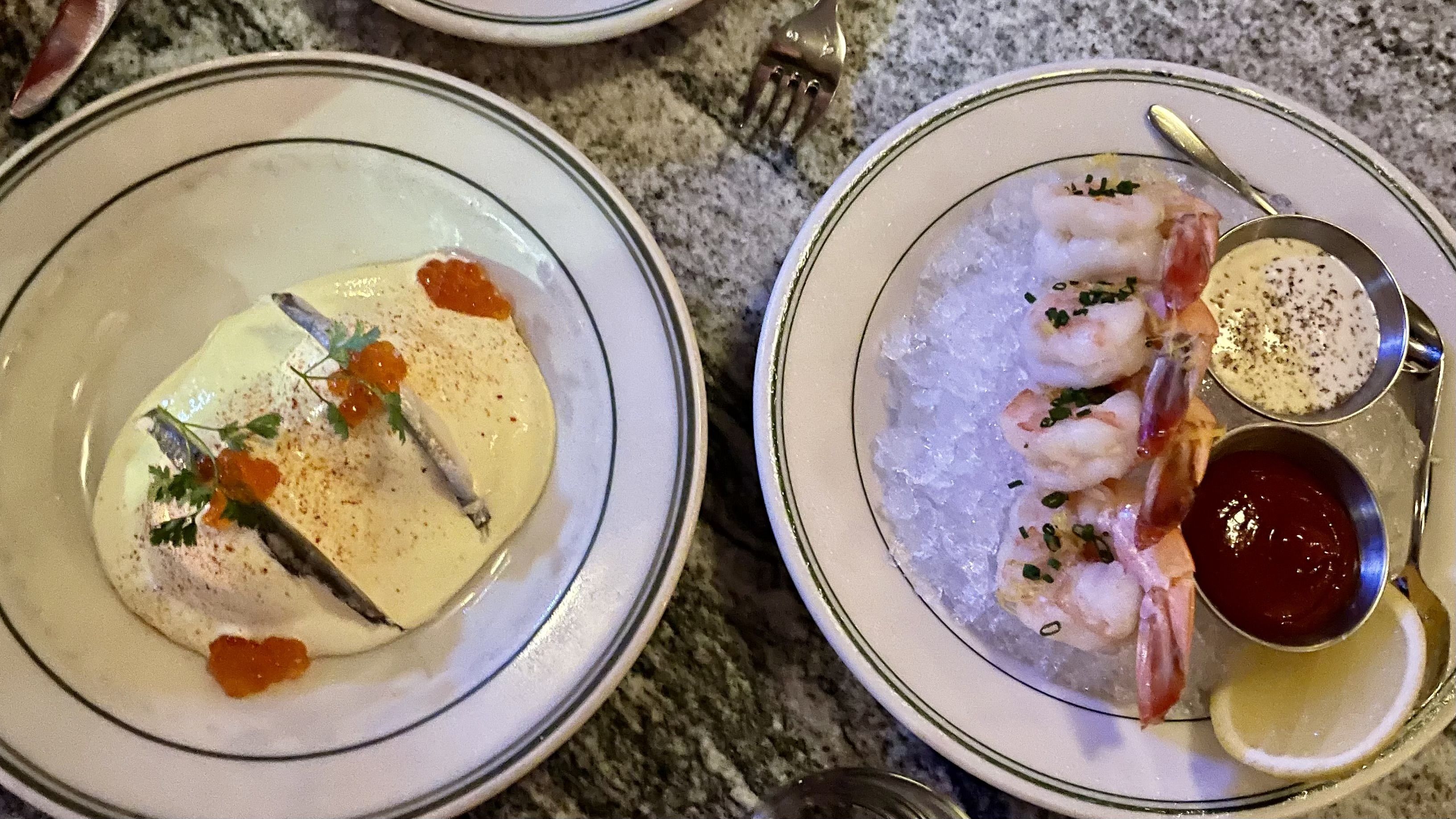 Two plates of seafood: left shows a pale creamy fish dish topped with orange roe and herbs; right features shrimp on crushed ice with lemon, a red dipping sauce, and a small mustard sauce.