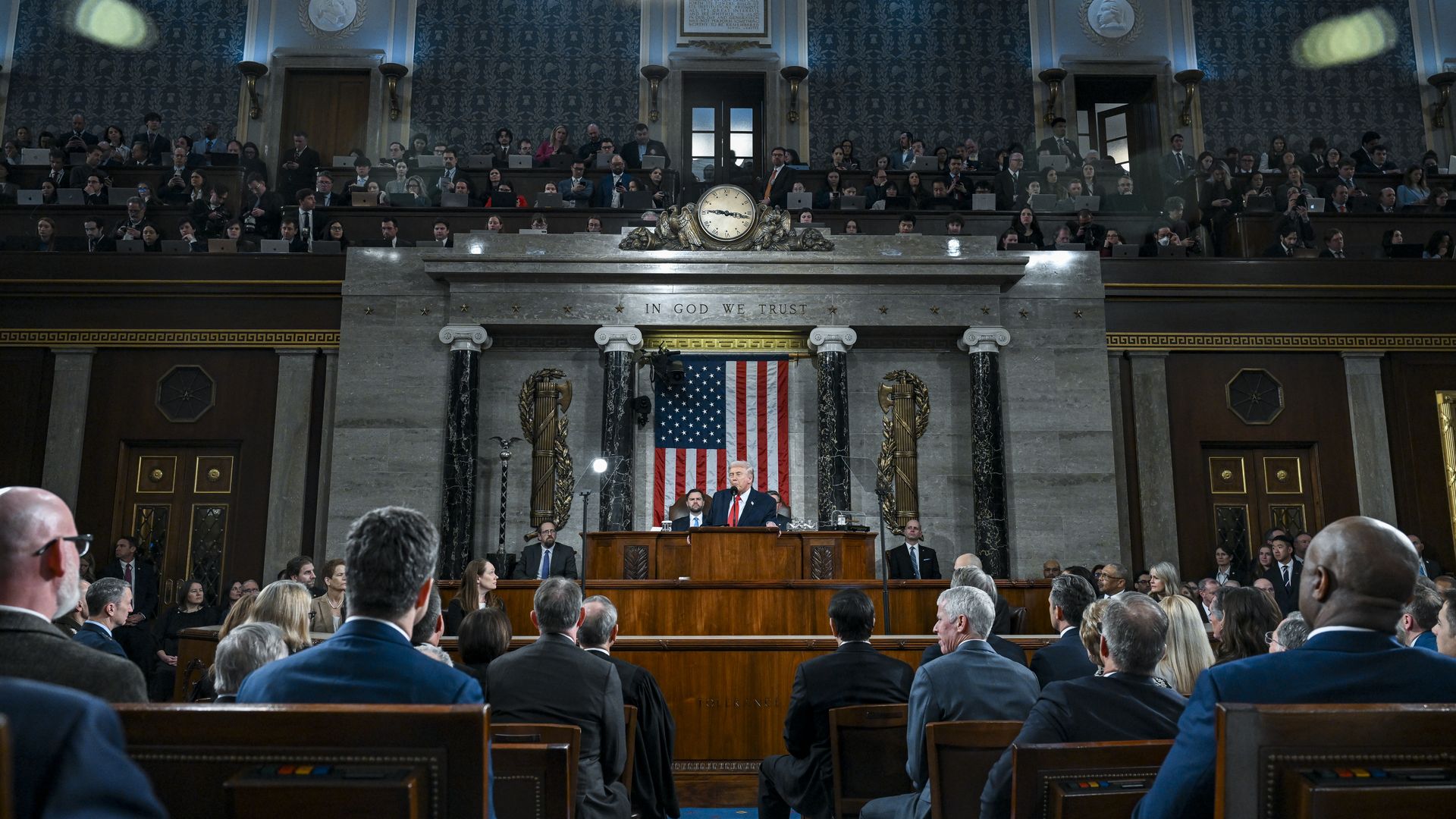 Speaker at a podium in a large chamber with an American flag and the phrase "IN GOD WE TRUST" above. Audience seated in front and multiple rows of people behind him.
