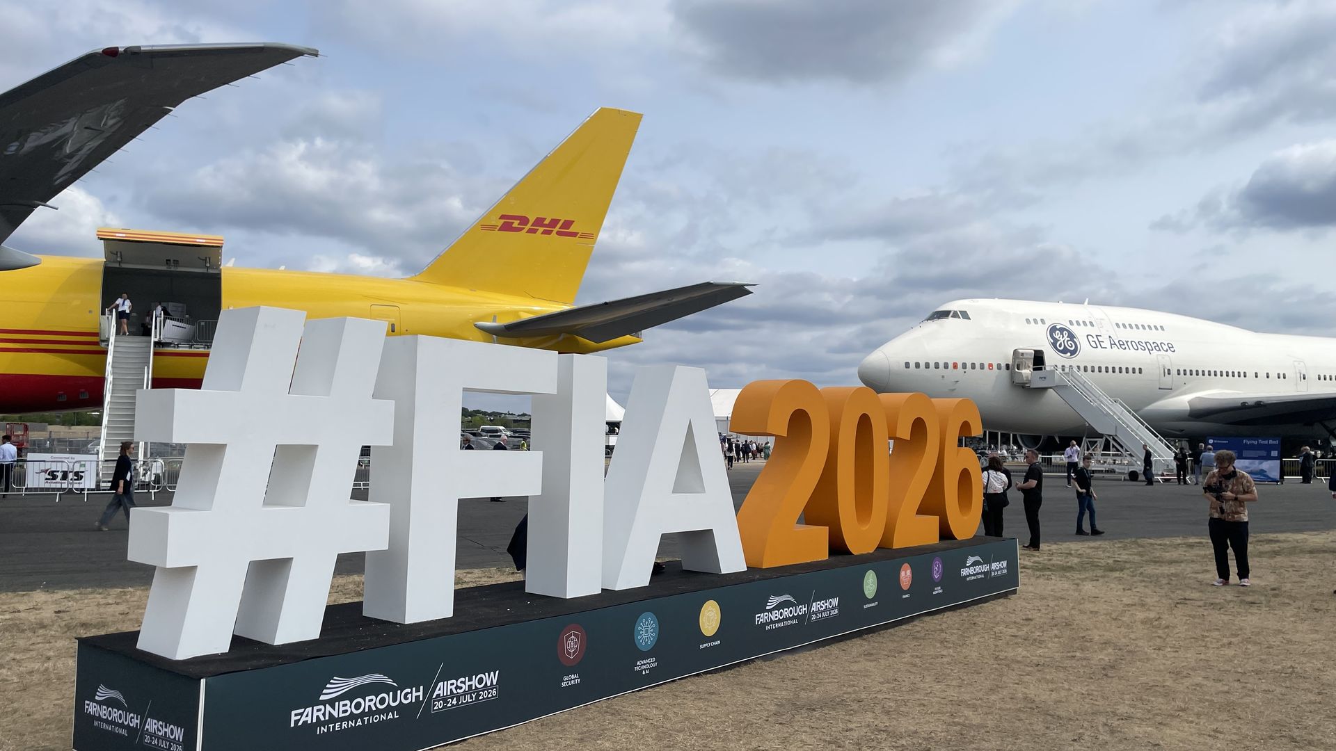 Outdoor airshow display featuring a bright yellow DHL cargo plane tail, a white GE Aerospace jet, a large white hashtag sculpture and orange #2020 text while visitors walk nearby under a cloudy sky.