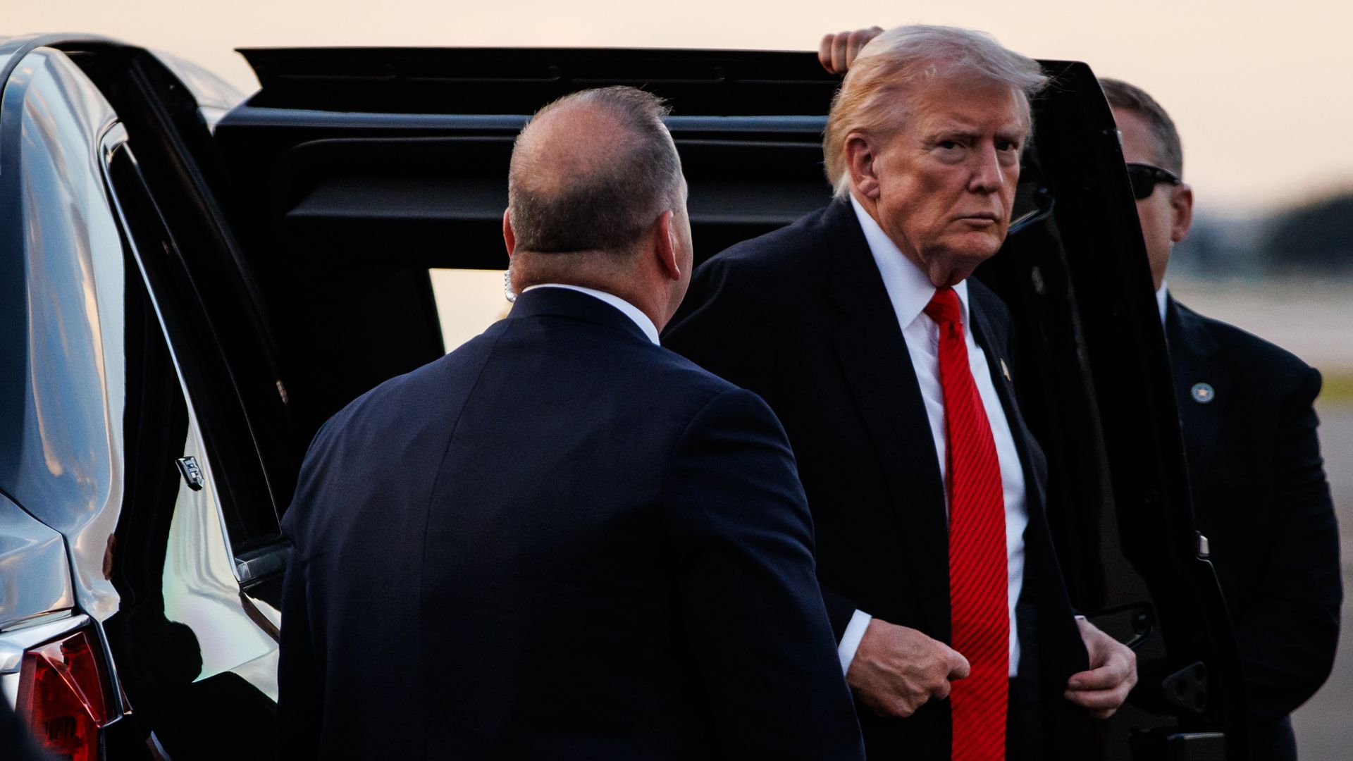 President Trump, wearing a navy jacket with a US flag pin at the top of his left lapel, white shirt and red tie, glares into the camera as he steps out of a car with two men in dark suits flanked either side of him.