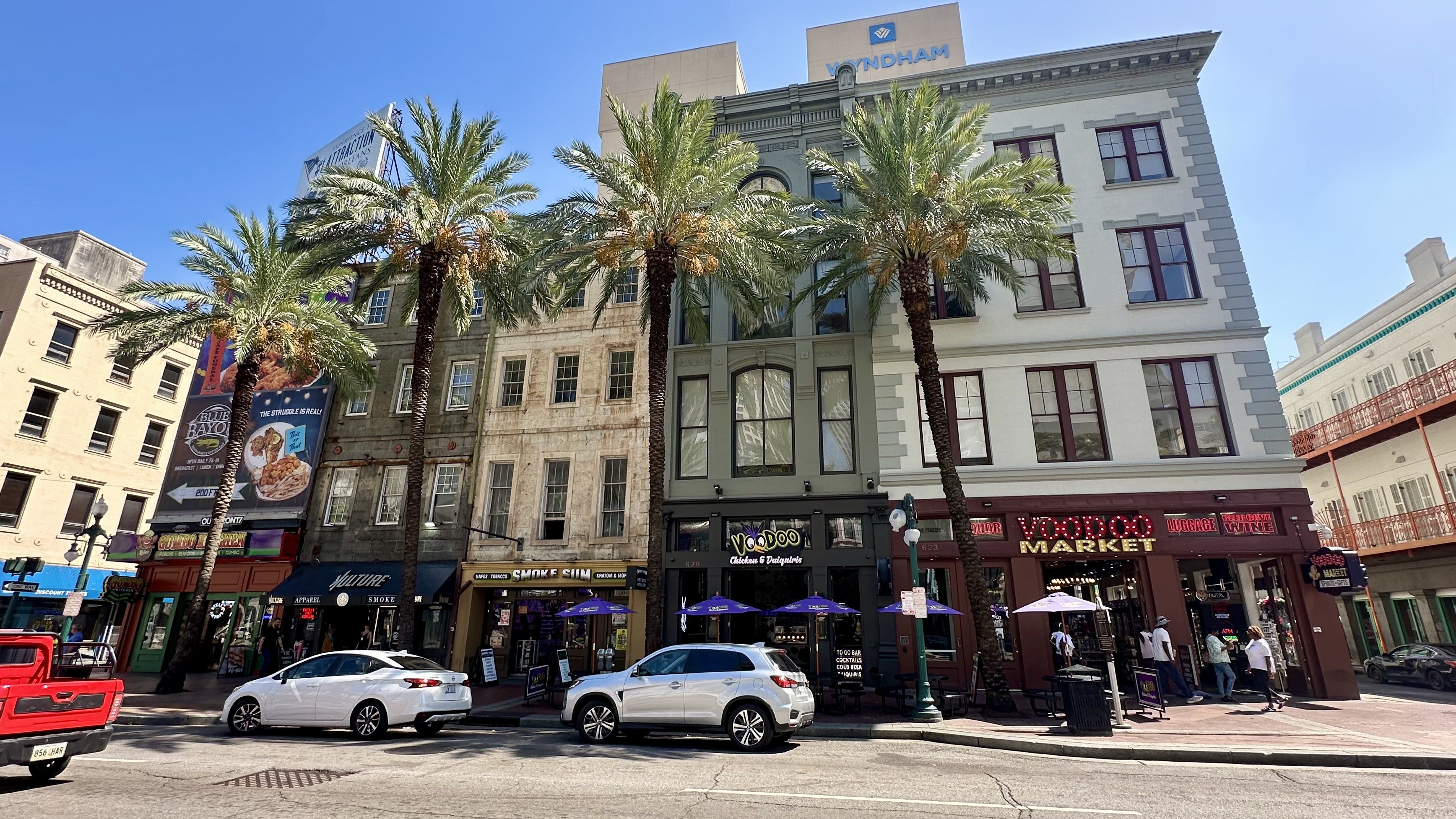 Street view of a sunny city block with palm trees and various storefronts including Voodoo Chicken & Daiquiris and Voodoo Market, with cars parked along the road and people walking.