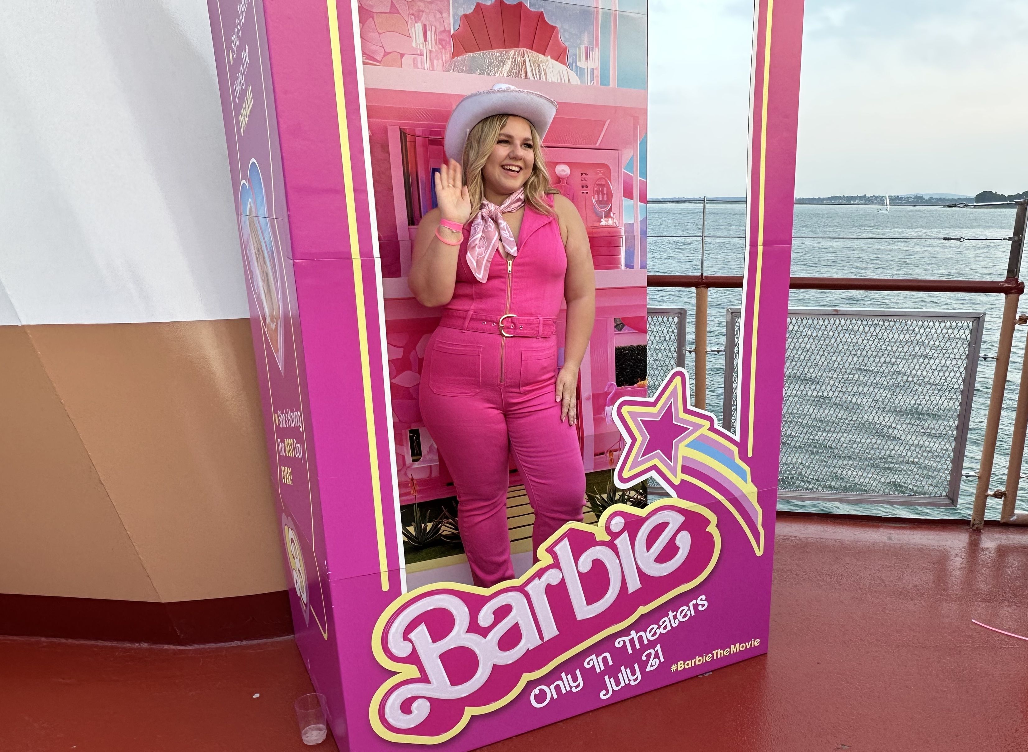 Katie Groves, a white blonde woman, poses for a photo in the life-size Barbie doll box. Katie's  wearing a pink jumpsuit and scarf and white cowboy hat.