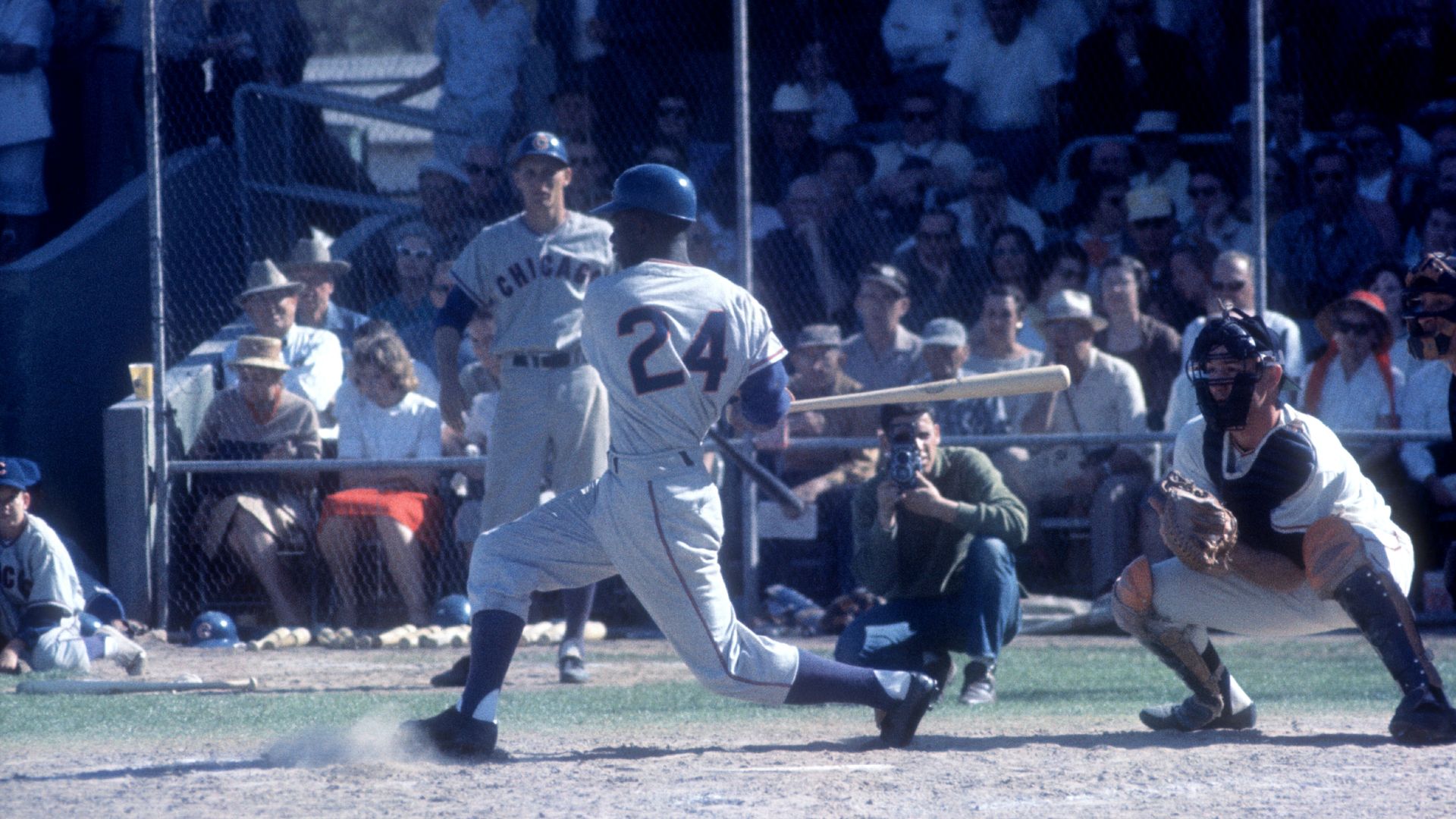 Baseball player wearing number 24 in gray uniform swings bat at pitch, catcher in black and white gear crouches behind him, with crowd and photographer in the background.