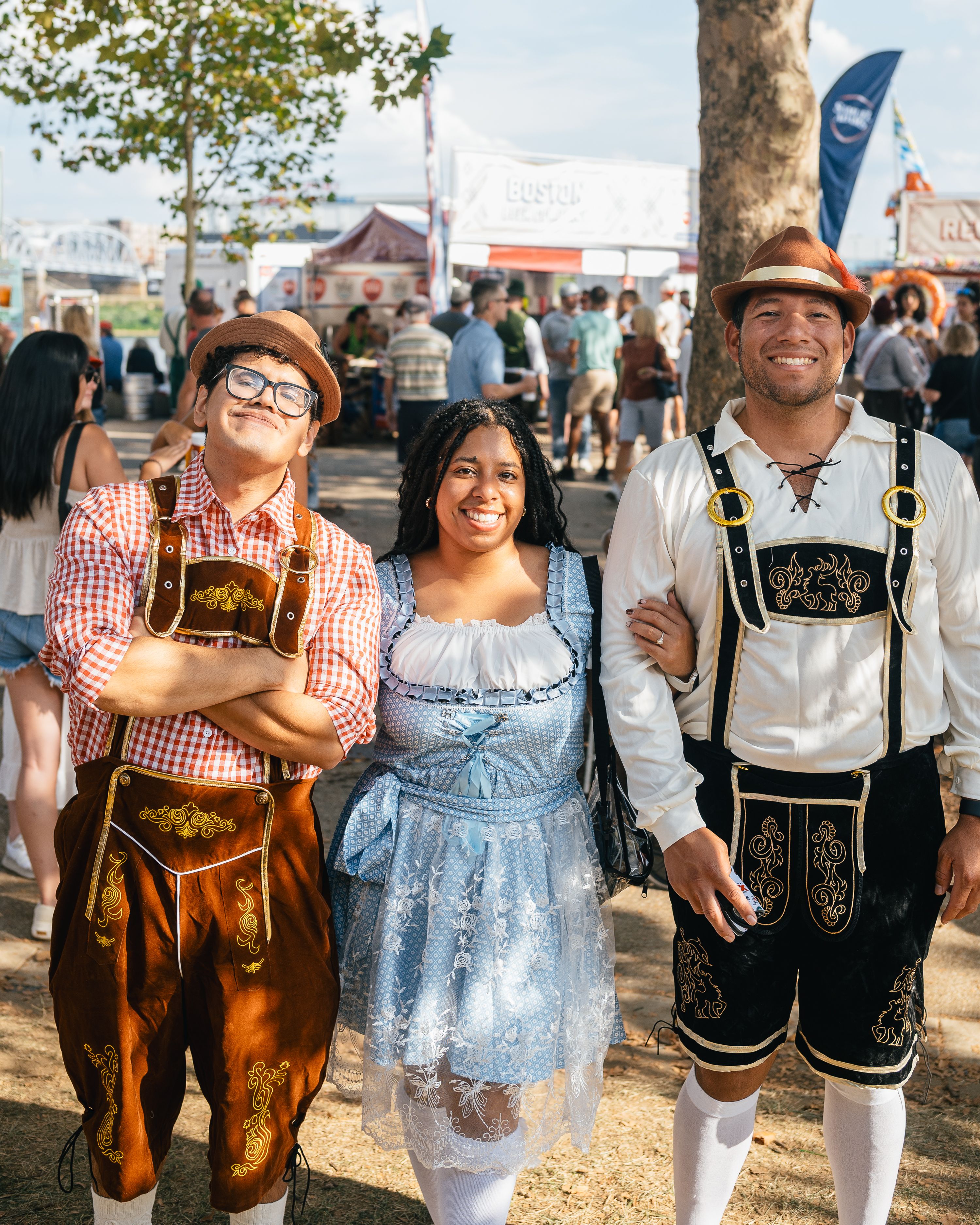 Three people in Bavarian outfits pose and smile at an outdoor festival; two men wear brown and black lederhosen and a woman wears a blue dirndl, with stalls and crowd behind them on a sunny day.