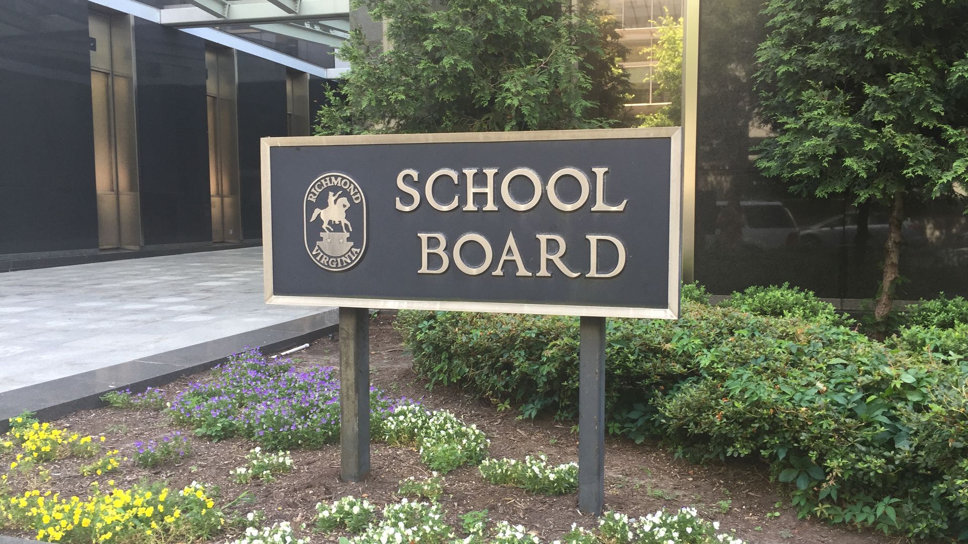 Black and gold sign for Richmond, Virginia School Board surrounded by green shrubs and colorful flowers outside a building with glass and stone walls.