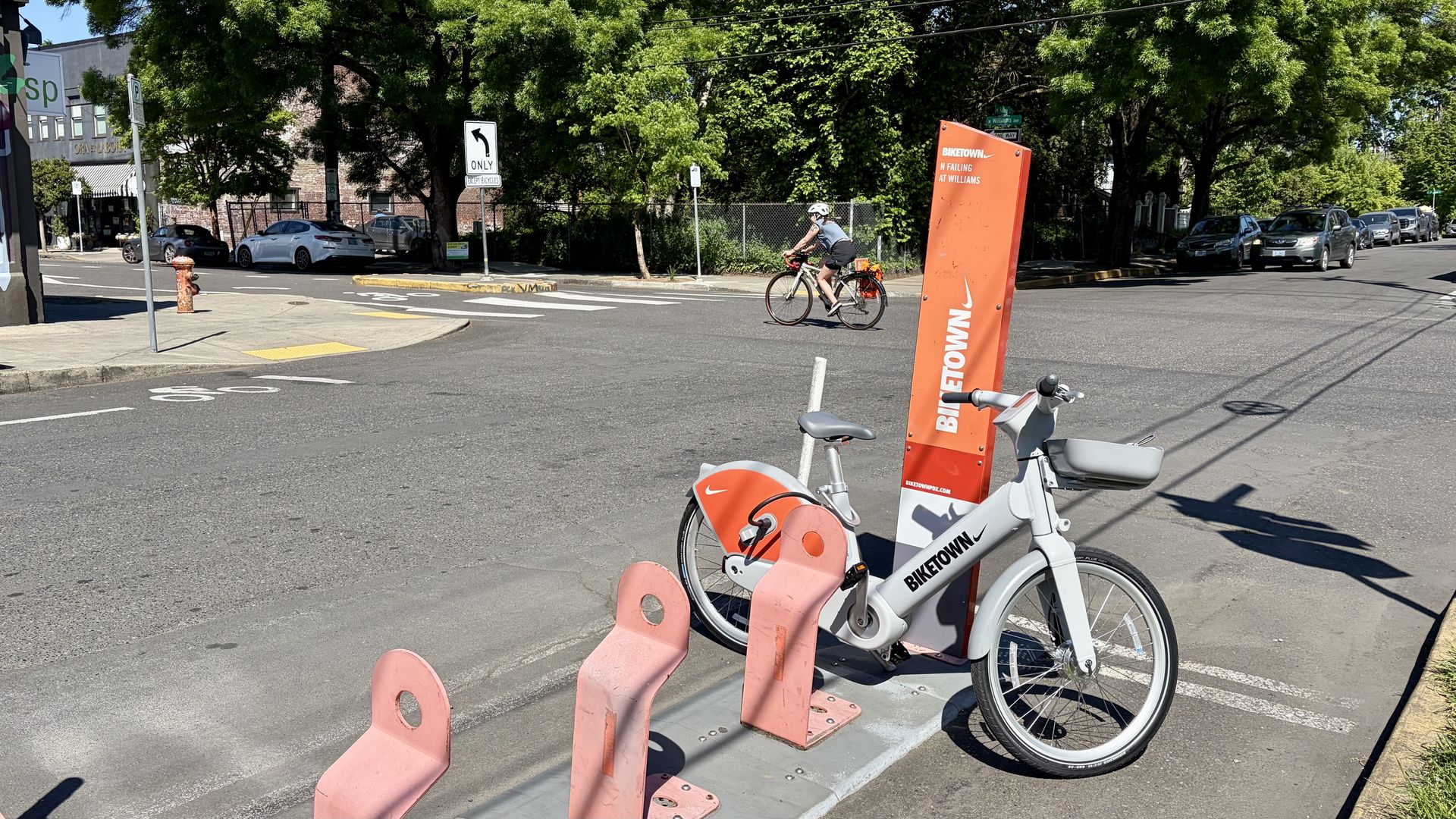Bright street scene with an orange BIKETOWN bike-share stand in the foreground, two bikes at the rack, a cyclist crossing the crosswalk, and a line of cars with green trees above.