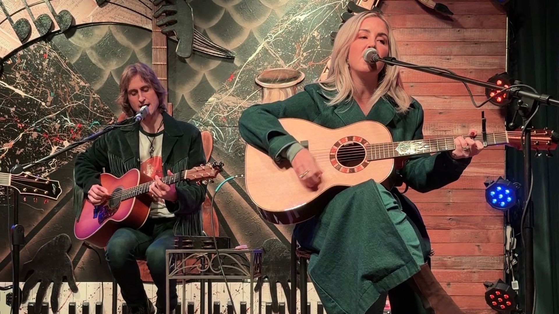 Live at Dave's, a blonde singer with an acoustic guitar performs at the mic while a guitarist with a purple guitar sits left; colorful lights and a music-backed mural on stage.