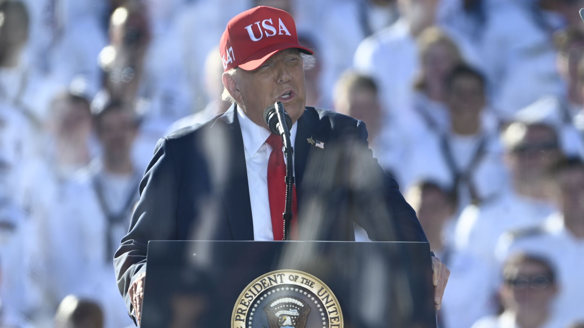 President Trump, wearing a red cap with white "USA" writing and a navy jacket with a US flag, white shirt and red tie, speaks into a mic in front of sailors in white uniform at a celebration for the Navy's 250th anniversary at Naval Station Norfolk in Norfolk, Virginia, on Sunday. 