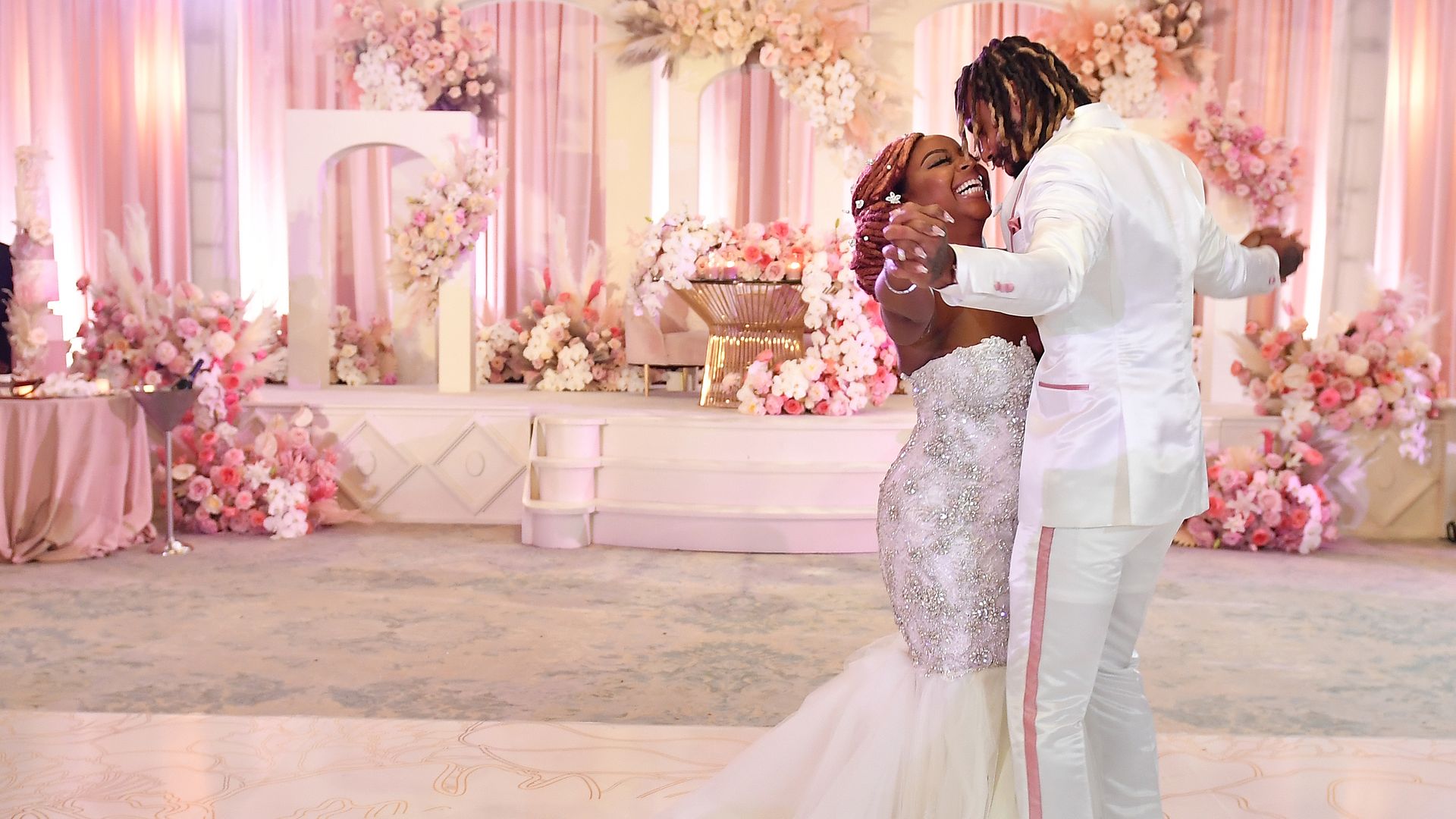 A bride wearing a white flowing dress and husband in a white tuxedo smile while sharing their first dance in a white and light-pink themed wedding