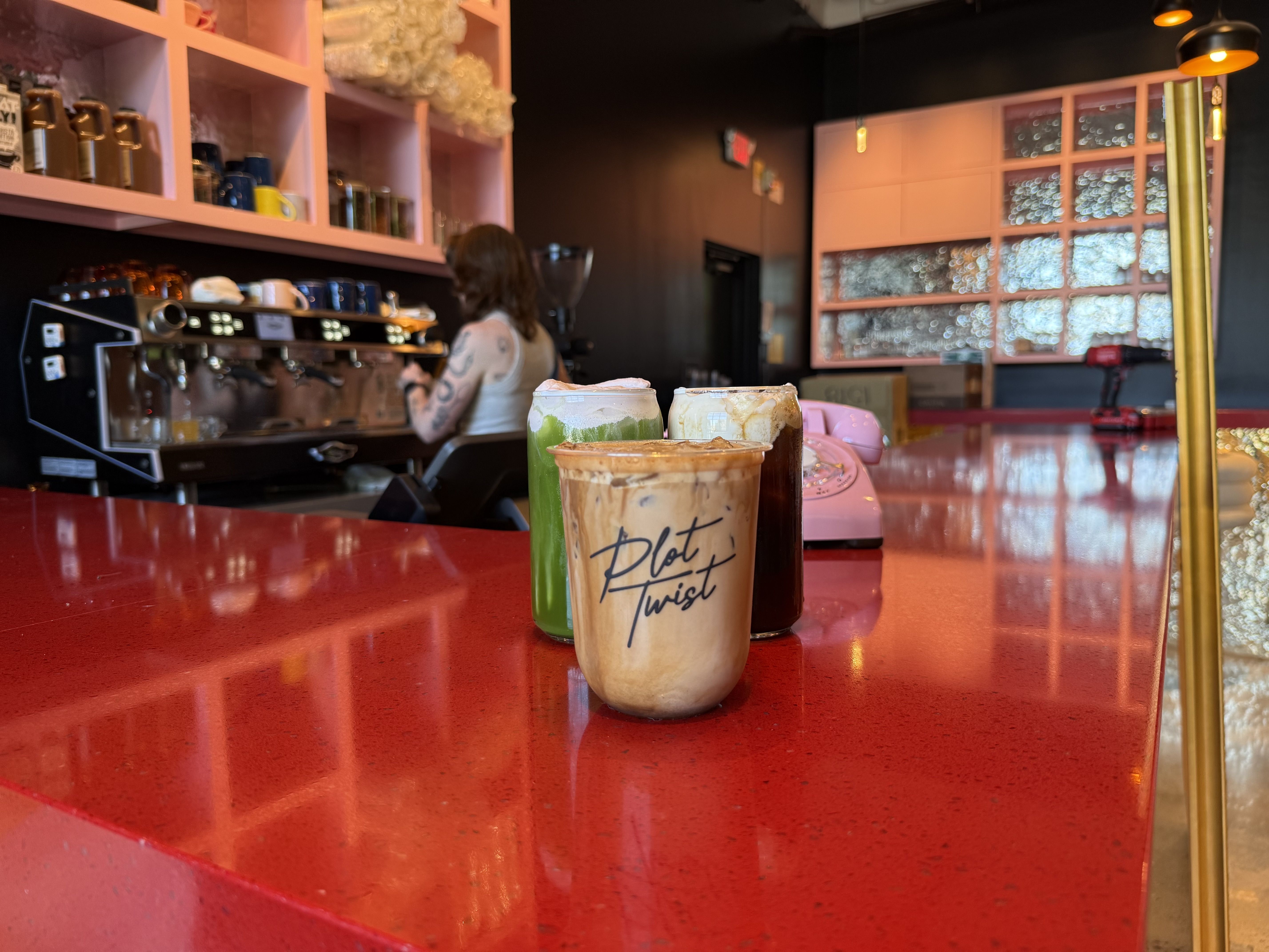 Three frothy drinks on a glossy red bar counter in a cozy cafe. Foreground cup reads Plot Twist. A barista works at an espresso machine in the background, with pink shelves and a retro phone nearby.