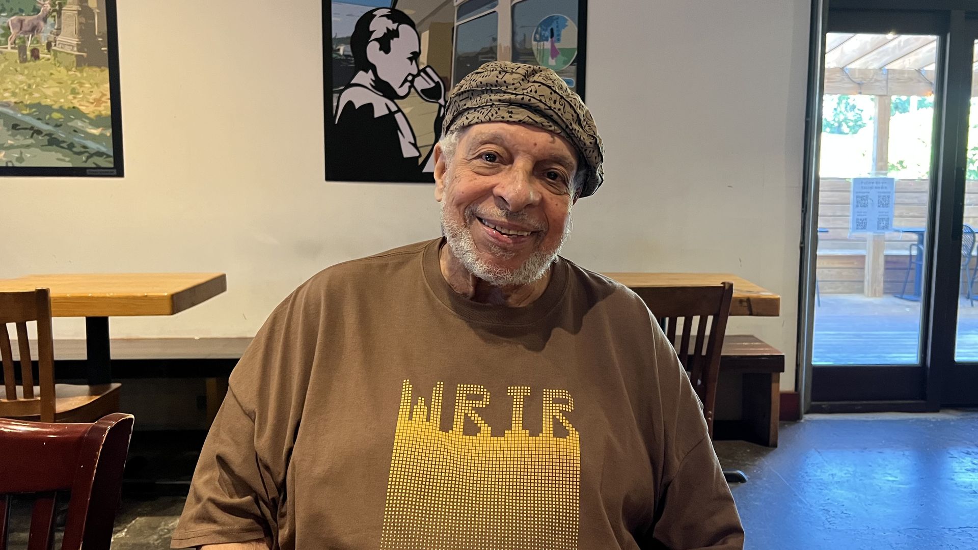 Smiling elderly man with a beard wearing a brown WRIR shirt and patterned cap, sitting in a café with wooden tables, chairs, and posters on the wall behind him.