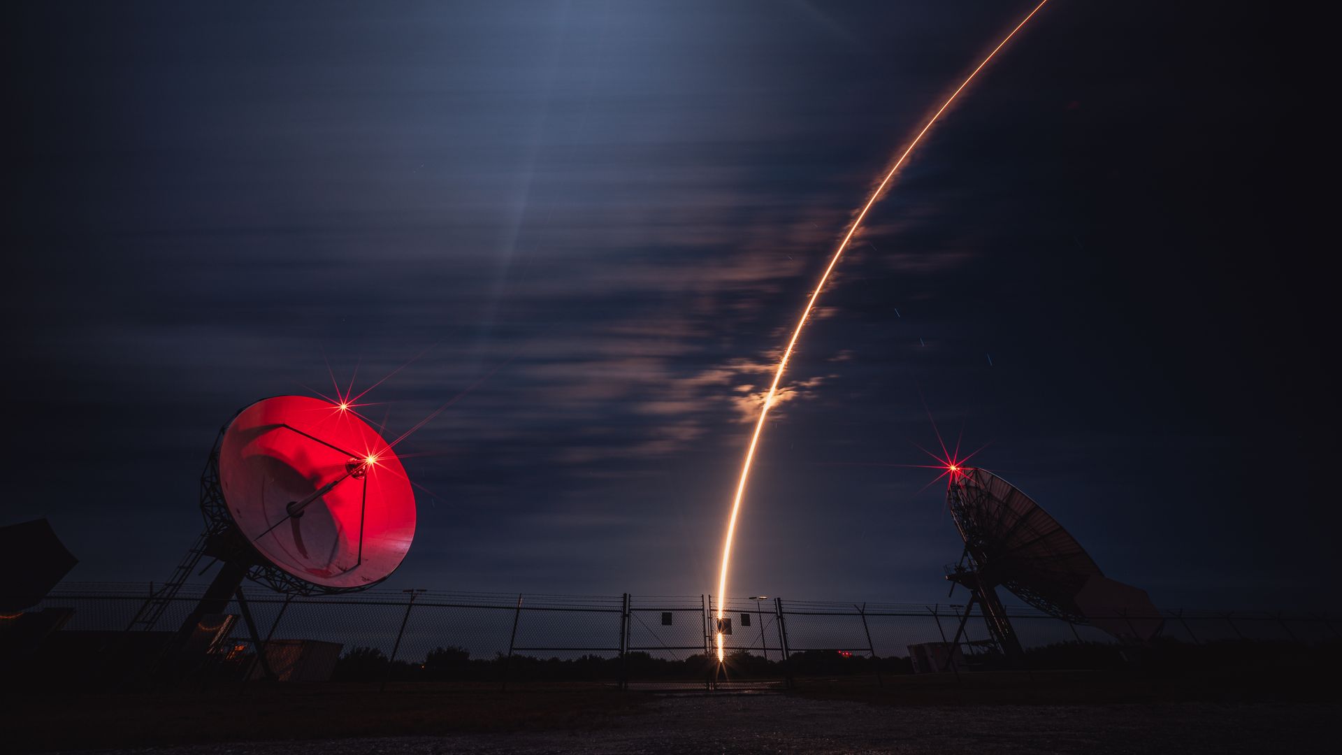 Nighttime scene with two large satellite dishes emitting red light and a bright orange arc of a rocket launch streaking across a cloudy sky above a fenced area.