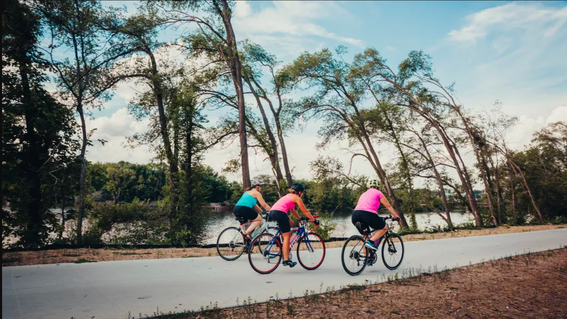 In this image, three bicyclists in brightly colored shirts and black shorts bike down a wooded street.