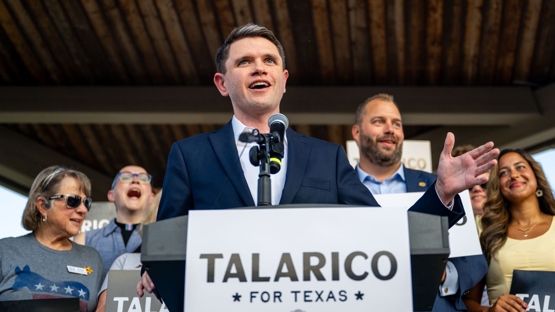 A man in a navy suit speaks at a podium labeled "TALARICO FOR TEXAS" with a microphone. Behind him, supporters cheer, including a woman in sunglasses and a smiling man, under a wooden ceiling.