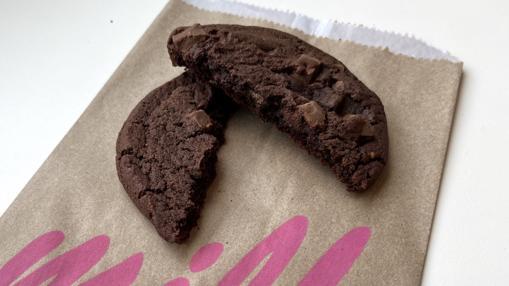 A chocolate cookie in two halves on top of a brown bag with the pink lettering milk.
