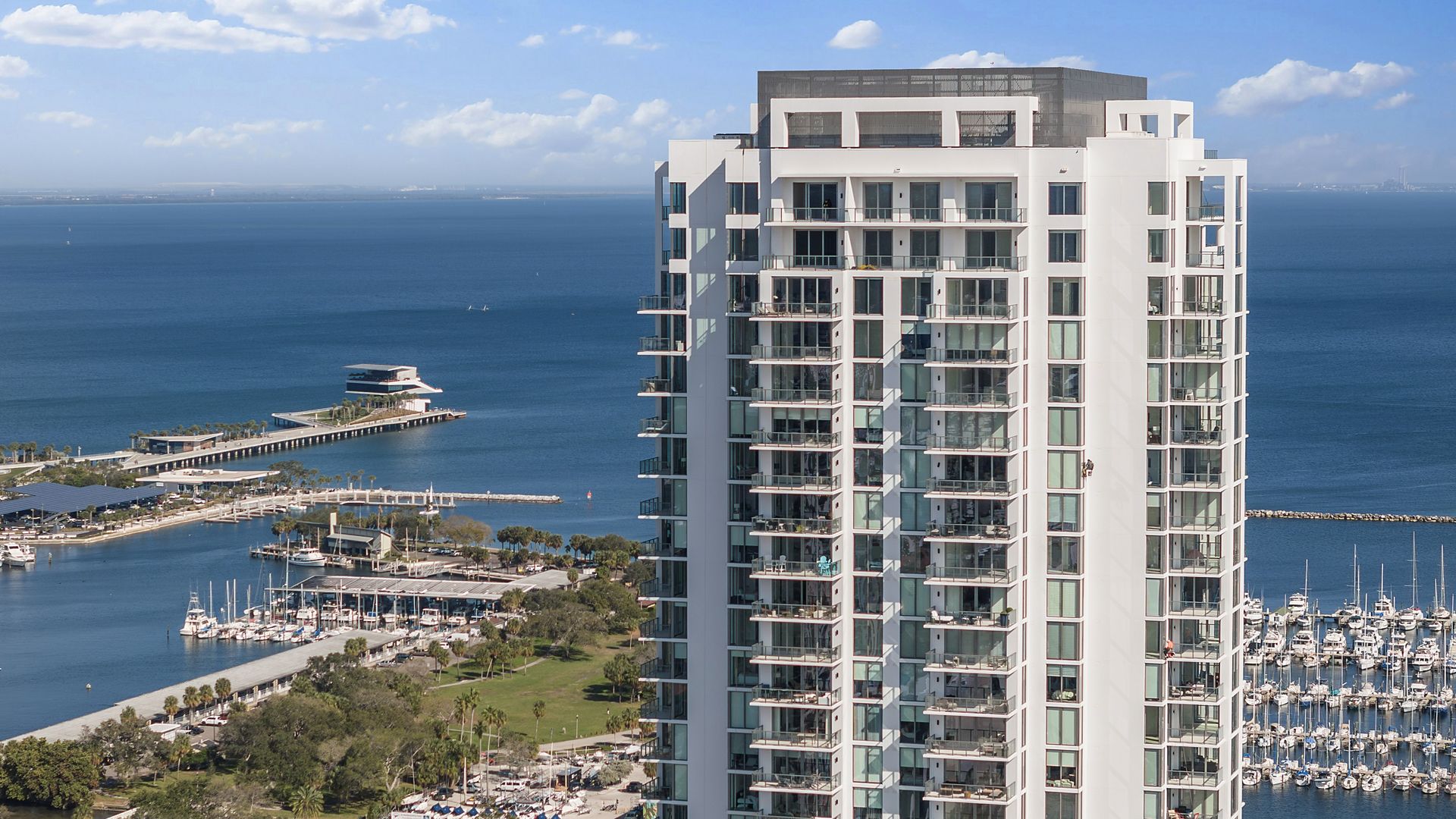 The top dozen floors of a high-rise condominium building with a view of water, a pier and a marina. The building is white and gray with lots of glass. It's very modern-looking.