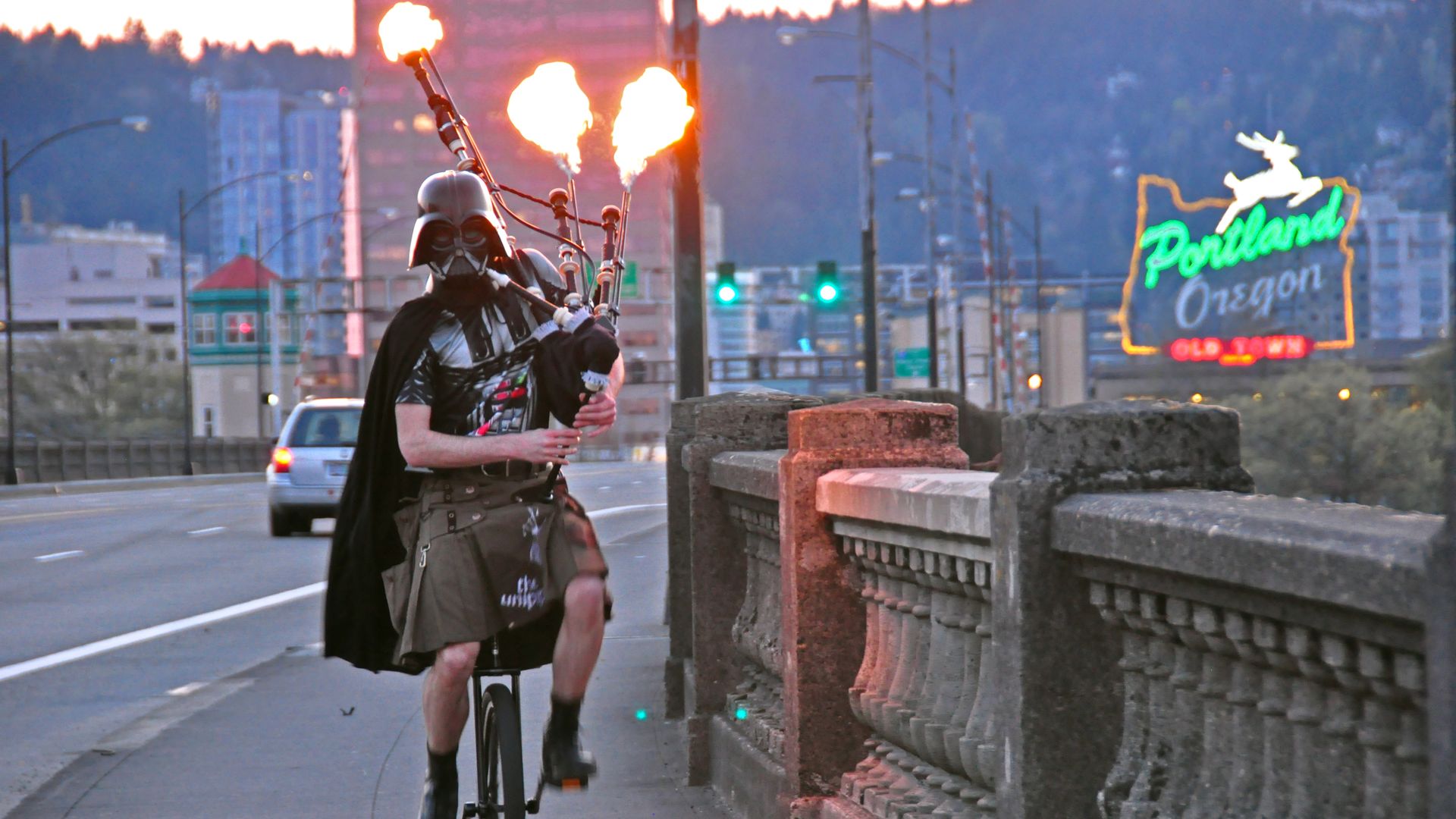 A man wearing a black Darth Vader mask and playing bagpipes with flames coming out of them unicycles across a bridge with a neon sign reading Portland, Oregon in the background.