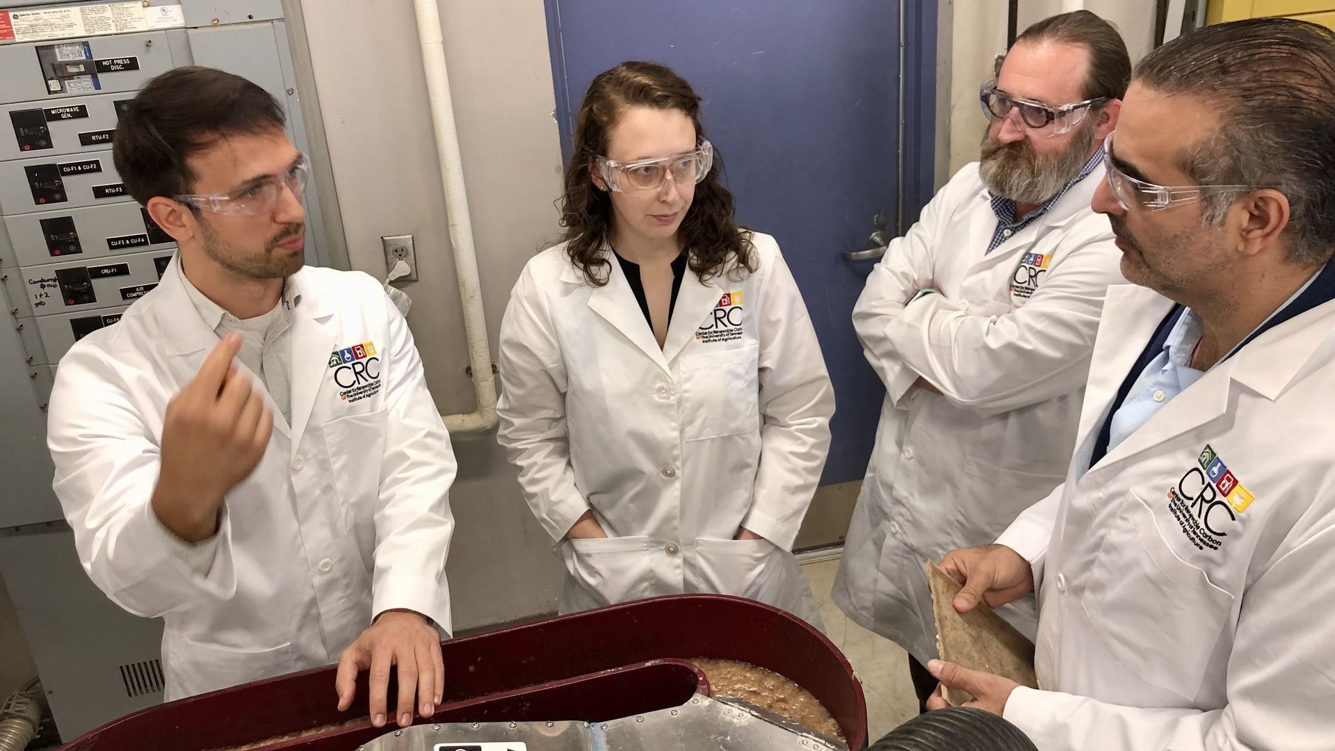 Four researchers in white lab coats and safety goggles examine a brown slurry in a large red hopper in an industrial lab, with control panels on the left and a blue door behind.