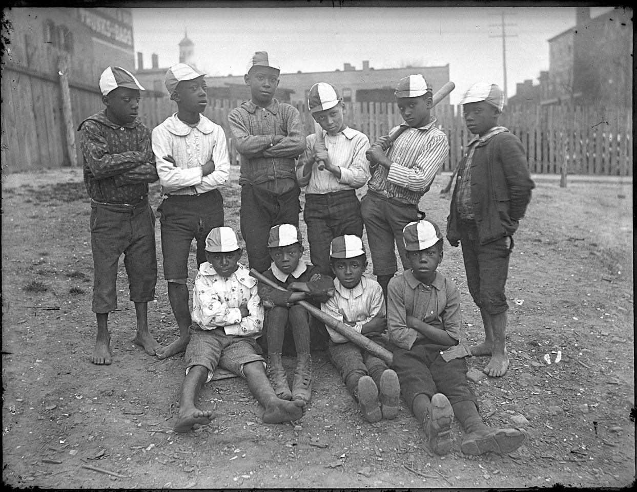 Glass plate negative showing a baseball team of young African-American boys; ten boys pose outside in an empty lot; four boys sit on ground; six boys stand behind them; all wear baseball caps; three hold baseball bats.