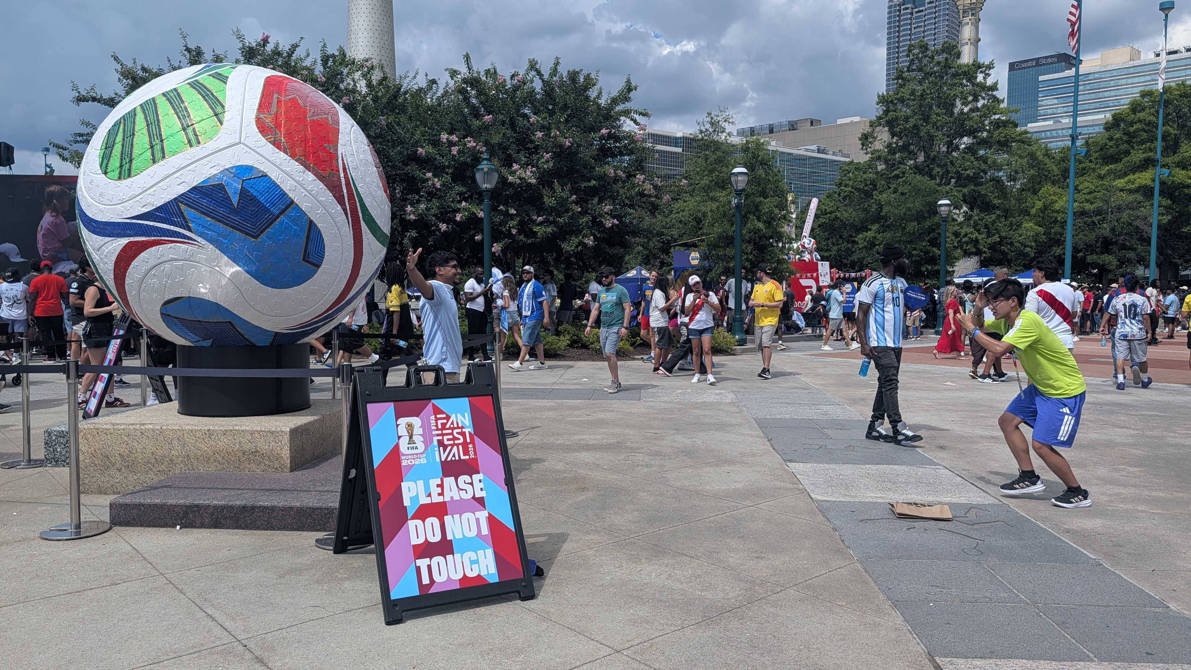 Large colorful soccer ball sculpture on a pedestal at an outdoor festival. A bright PLEASE DO NOT TOUCH sign in the foreground; crowd, trees, and city skyline in the background.