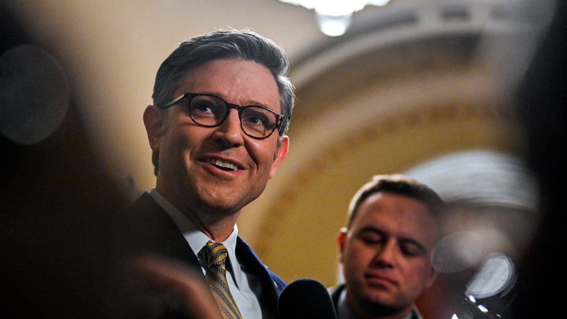 House Speaker Mike Johnson, flanked by reporters standing in front of a yellow wall.
